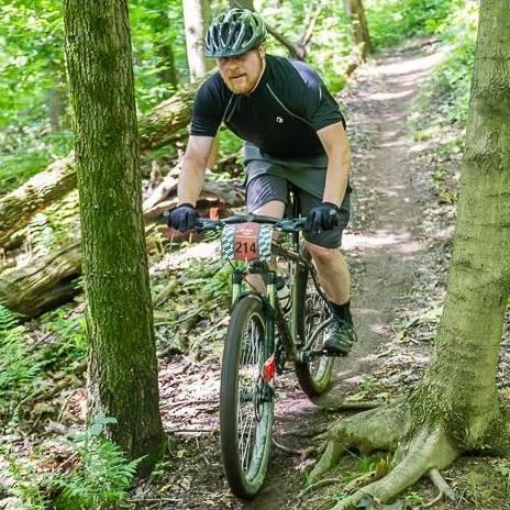 A man riding a mountain bike on a dirt trail surrounded by green trees and foliage. He is wearing a helmet, a short-sleeve shirt, and shorts, and has a race number attached to his bike. The trail is narrow and features roots and fallen branches. Brady's Run County Park mountain bike trail.