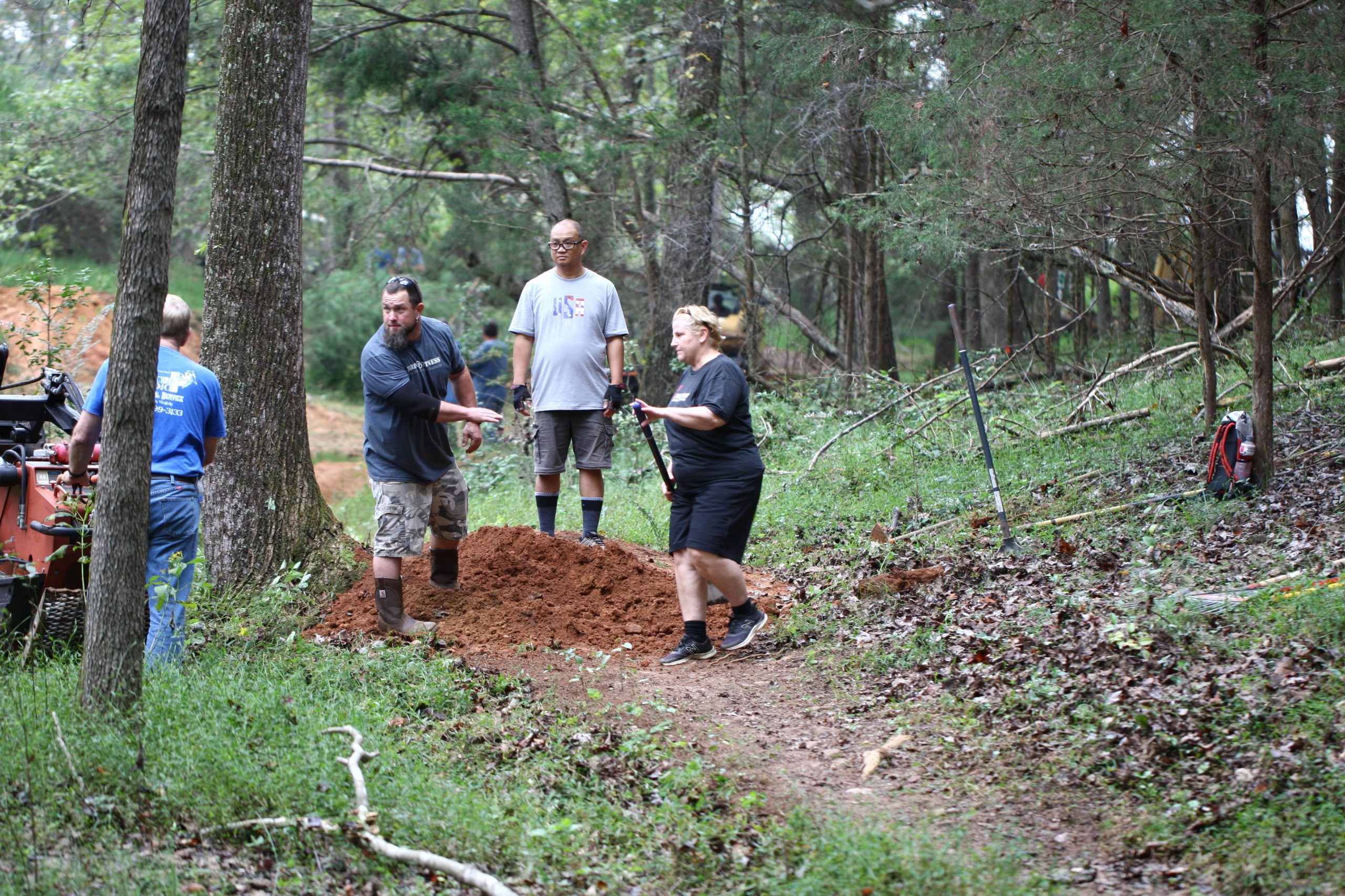 Four individuals are working together outdoors in a wooded area. They are engaged in digging and moving dirt, with one person using a shovel and another gesturing as they communicate. The scene is surrounded by trees and greenery, with some tools and equipment visible nearby. The group appears focused on their task in this collaborative effort. Mountain Laurel Trails mountain bike trail.