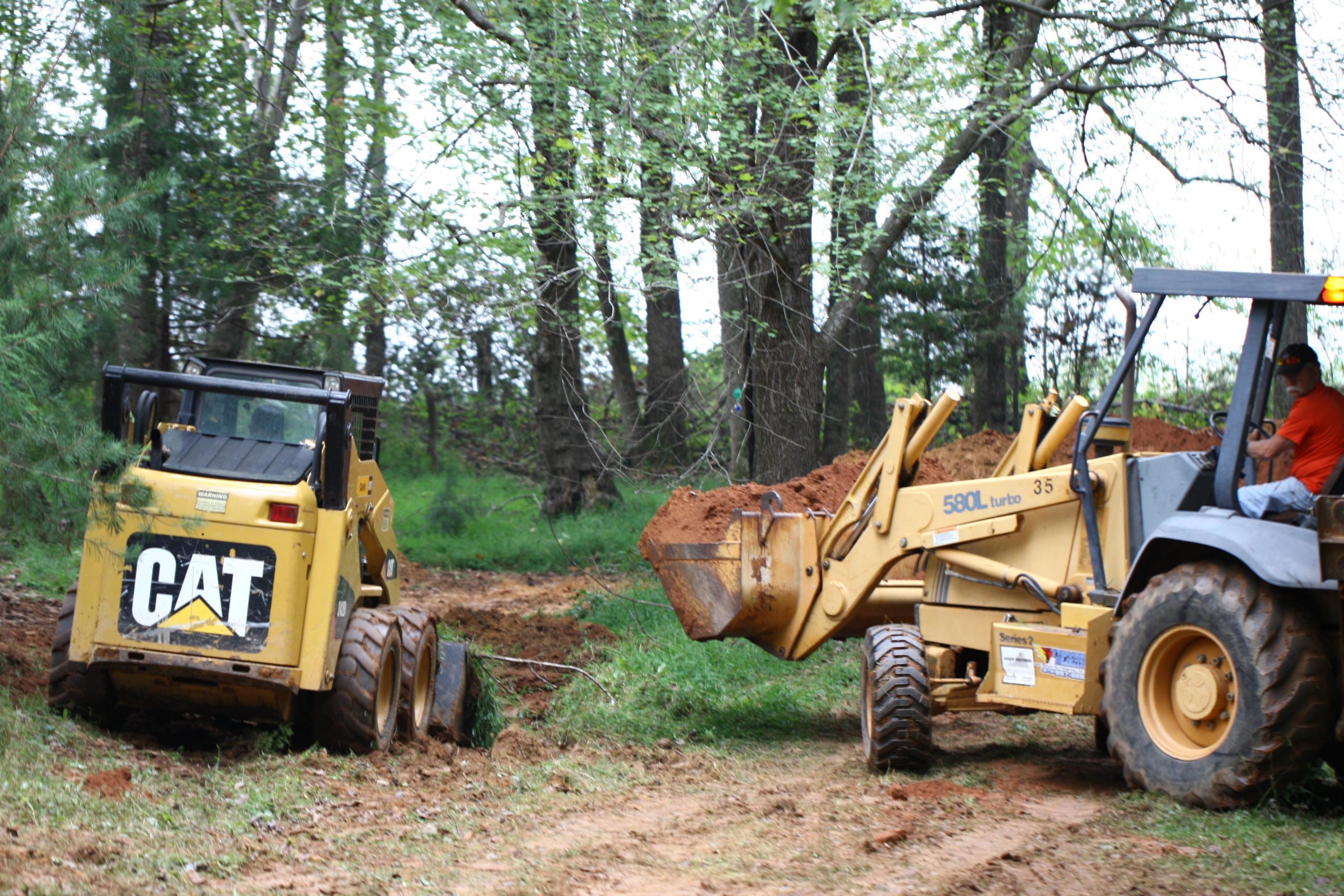 Two construction vehicles operating in a wooded area: a yellow skid steer loader marked with "CAT" on the back and a yellow backhoe loader, both moving dirt. The background features trees and greenery. Mountain Laurel Trails mountain bike trail.