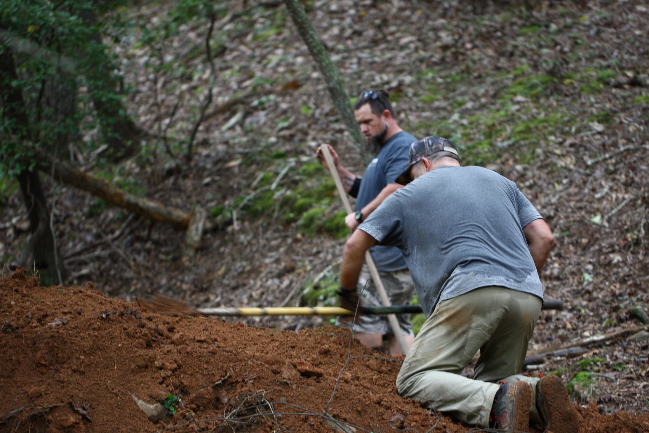 Two individuals are working outdoors on a sloped, earthy terrain. One person is kneeling and digging into the soil with a tool, while the other stands nearby, holding a shovel. The background features a forested area with trees and scattered leaves, suggesting an active outdoor project. Mountain Laurel Trails mountain bike trail.