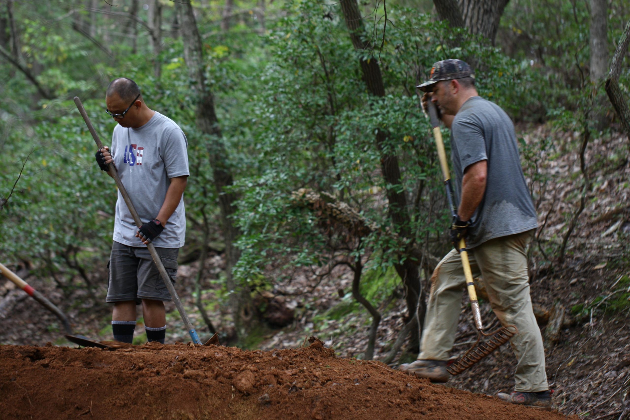 Two individuals are working in a wooded area, using gardening tools. One person is holding a rake, while the other is holding a long-handled tool. Both are dressed in casual clothing suitable for outdoor work. The ground around them is earthy and uneven, surrounded by green vegetation and trees in the background. Mountain Laurel Trails mountain bike trail.