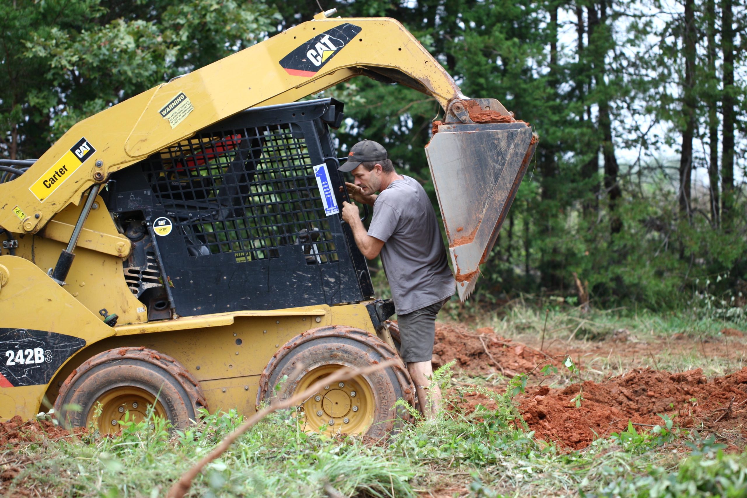 A person operating a yellow skid steer loader in a dirt field, leaning against the machine while inspecting the equipment. Surrounding vegetation includes green trees and sparse grass. Mountain Laurel Trails mountain bike trail.