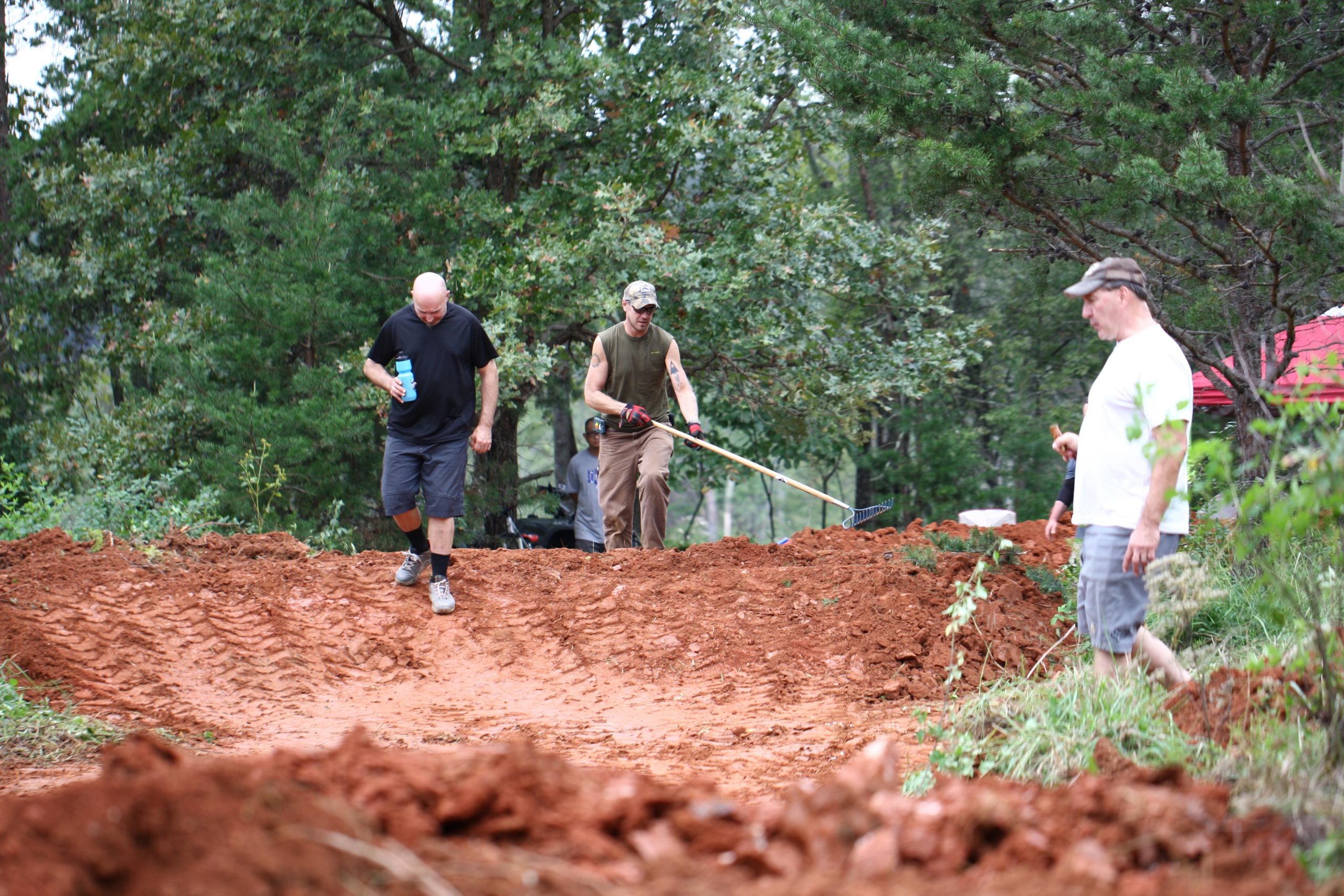 Three men work on a dirt trail surrounded by trees. One man in a black shirt walks with a water bottle, while another in an olive green shirt uses a rake to level the ground. A third man in a white shirt observes the work. The area has been recently disturbed, showing reddish-brown soil and tire tracks. Mountain Laurel Trails mountain bike trail.