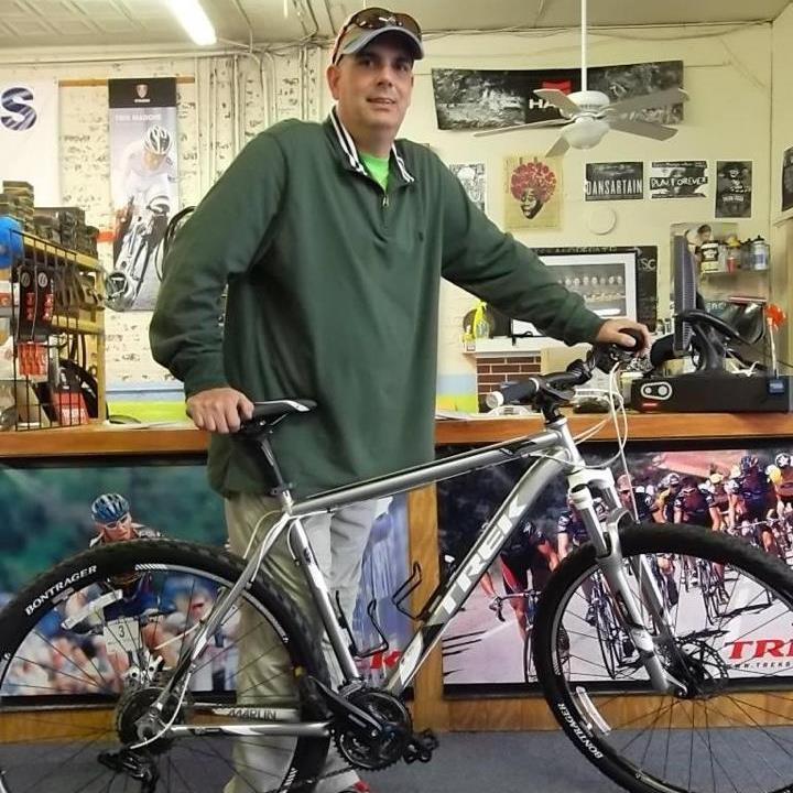 Trek Marlin: A man in a green long-sleeve shirt and a baseball cap stands beside a silver Trek bicycle, posing in a bike shop filled with cycling equipment and posters in the background.