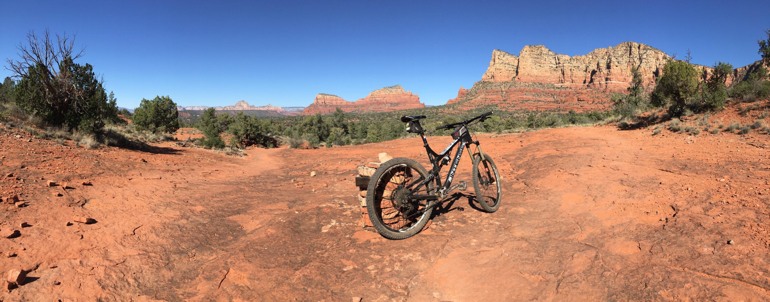 A mountain bike resting on a rocky, red dirt trail with green vegetation surrounding it, set against a backdrop of rugged rock formations under a clear blue sky. Bell Rock Area Trails mountain bike trail.