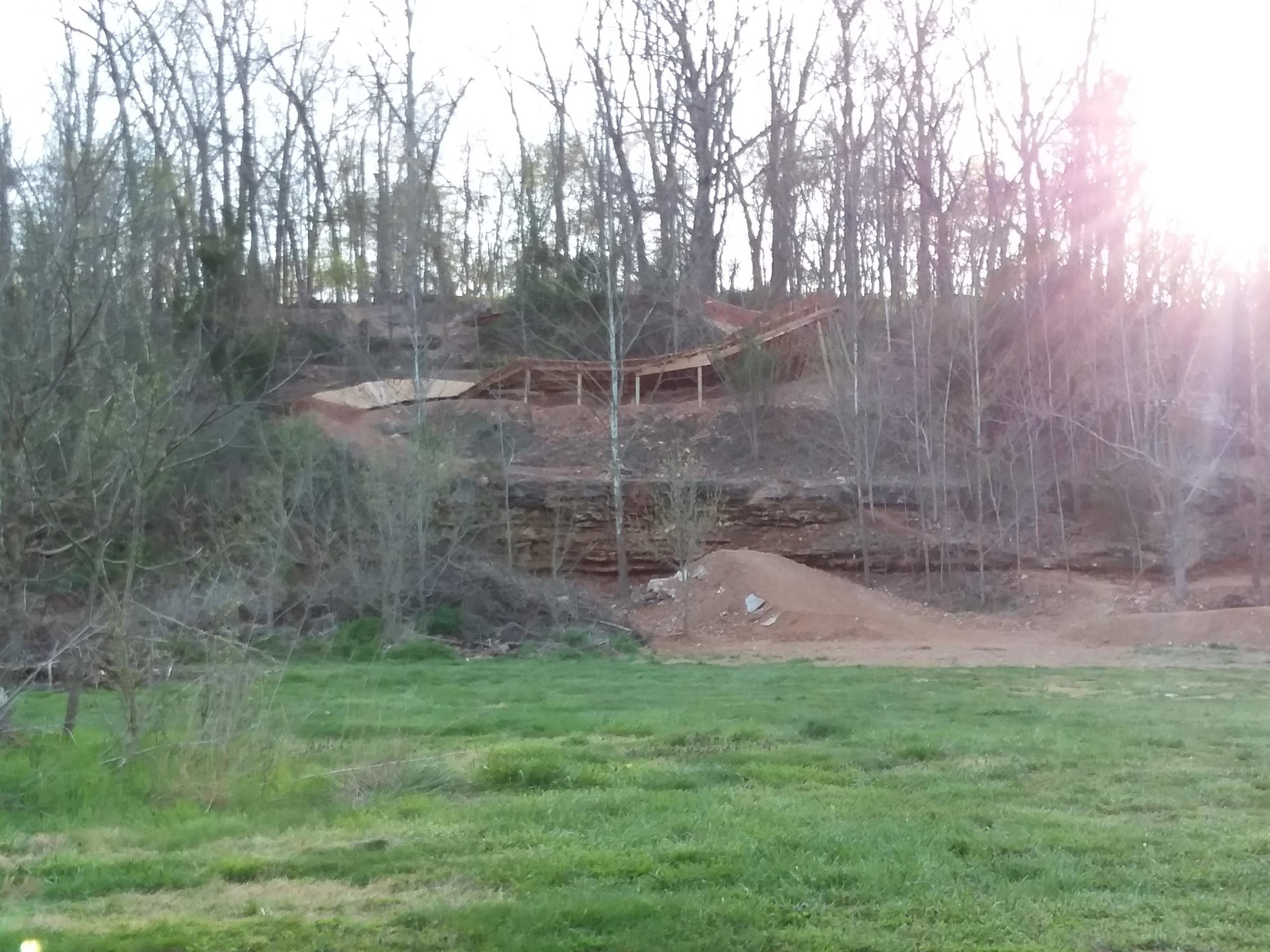 A scenic view of a wooded area showcasing a hillside with a dirt ramp and a wooden structure, surrounded by bare trees and green grass in the foreground. The sun is setting, creating a warm glow in the background. Slaughter Pen Trail mountain bike trail.