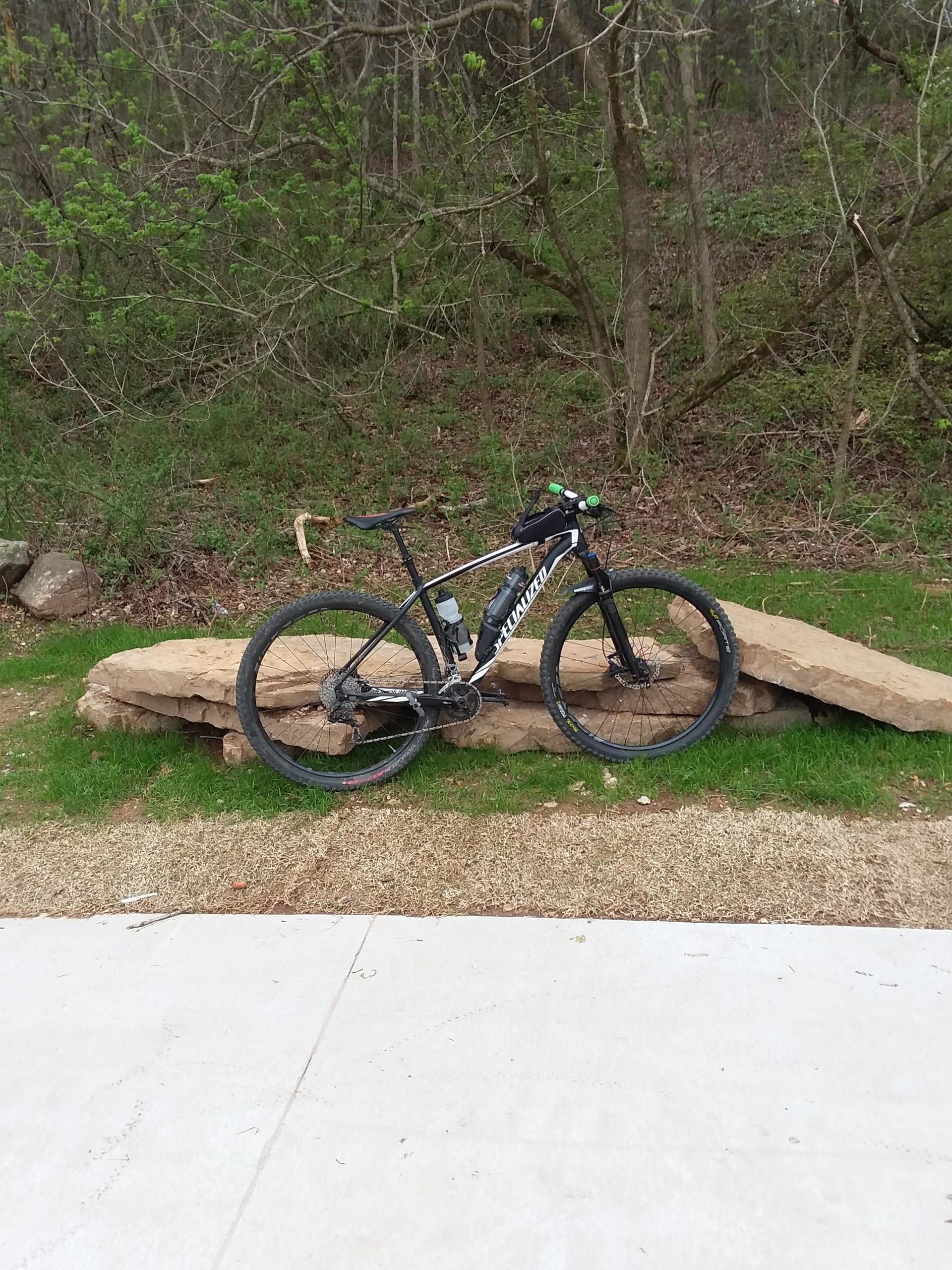 A black mountain bike with green accents is resting on a large rock, surrounded by greenery and trees in the background. The ground is a mix of grass and gravel, with a paved path in the foreground. Back 40 mountain bike trail.
