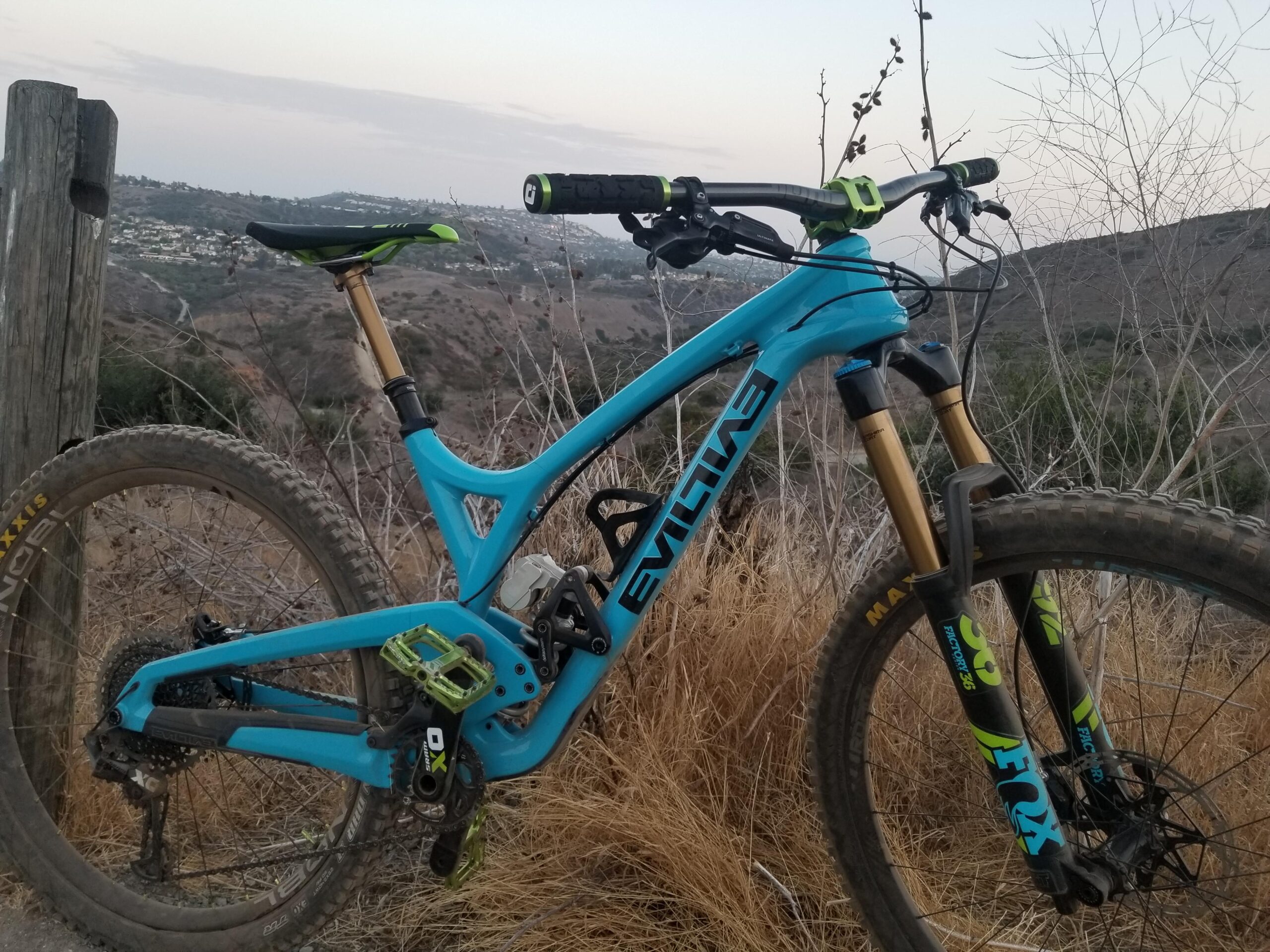 Evil Wreckoning: A close-up view of a blue mountain bike parked next to a wooden post, showcasing its suspension fork, pedals, and a background of rolling hills and sparse vegetation. The bike features branding and components from prominent brands, with a pathway visible in the distance. The scene is set during twilight with a soft, overcast sky.