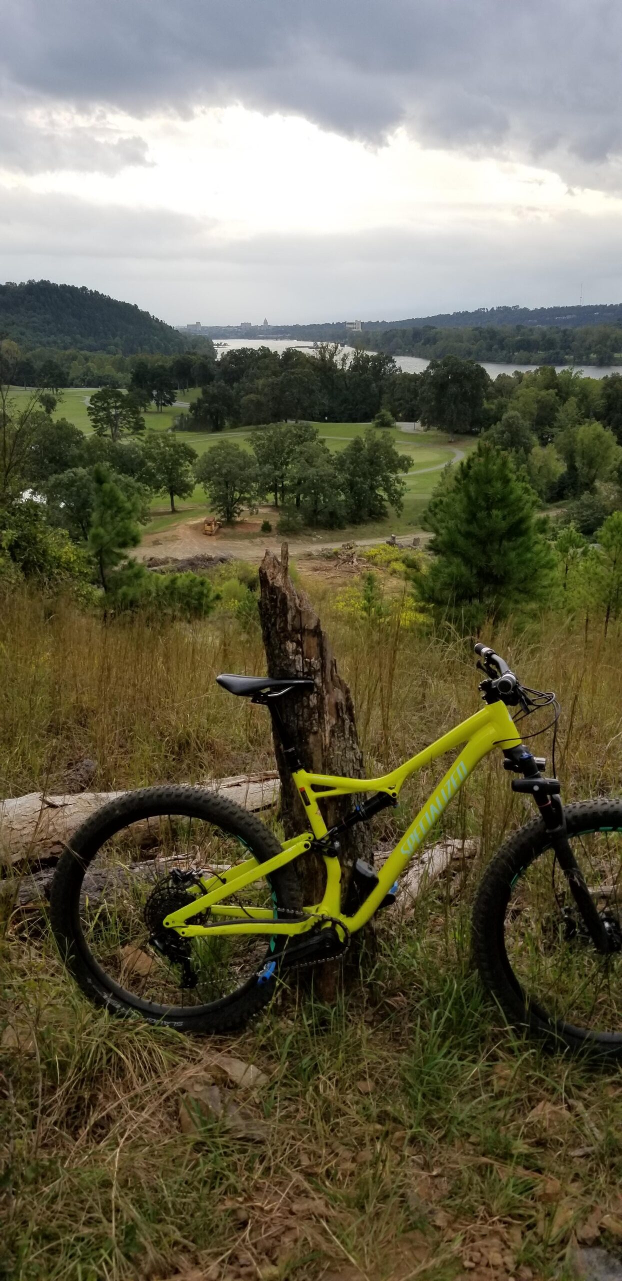 Specialized Stumpjumper FSR 6Fattie: A bright yellow mountain bike is leaned against a tree stump on a grassy hillside, overlooking a scenic landscape. In the background, a river stretches into the distance, bordered by trees and rolling hills under a cloudy sky.
