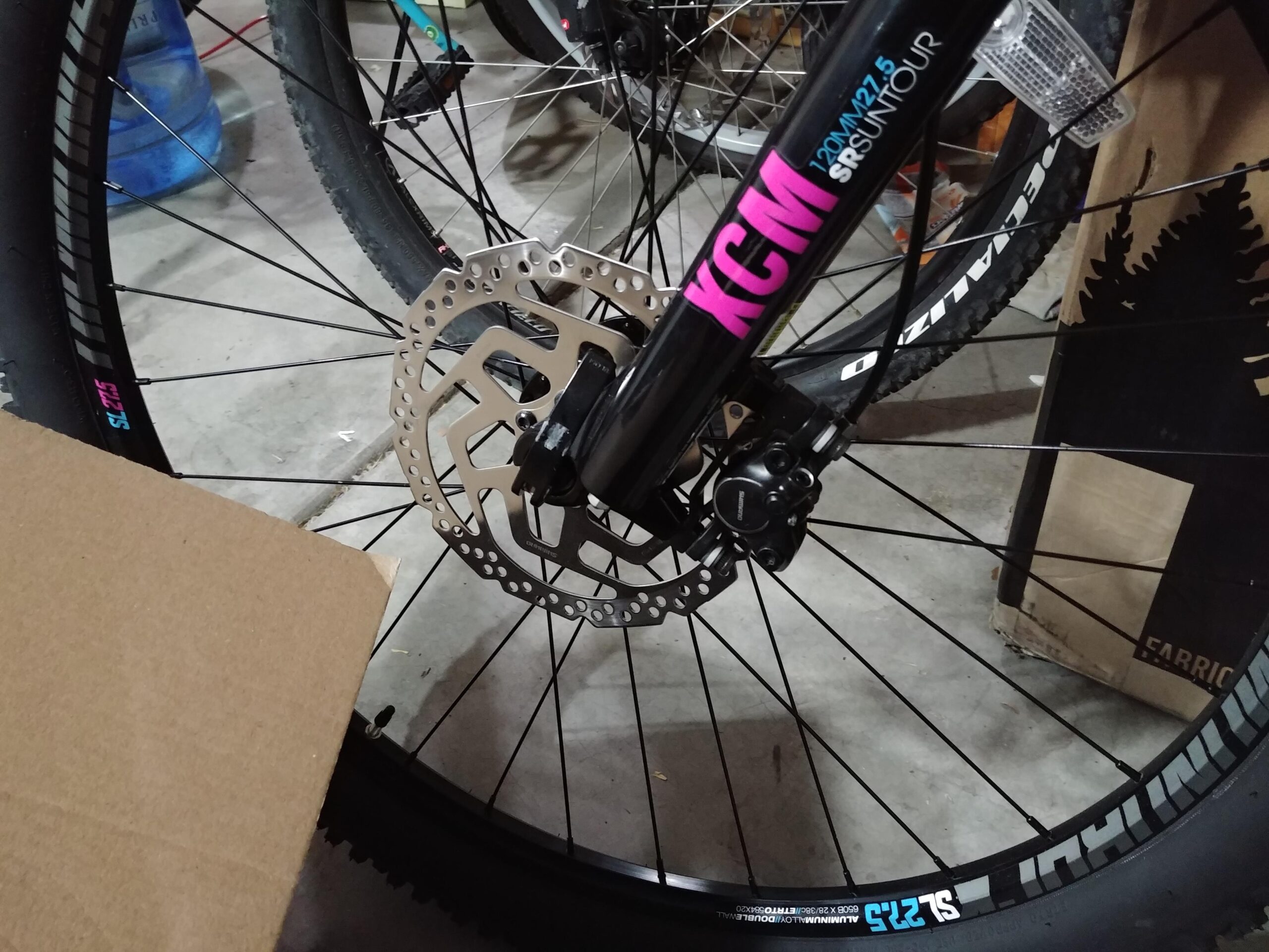 Diamondback Atroz 2 2018: A close-up view of a mountain bike's front wheel, showcasing the disc brake system and tire. The wheel features black spokes and a wide tire, while the bike's frame includes branding and specifications. A cardboard box is partially visible in the foreground, with a concrete surface contributing to an indoor setting.