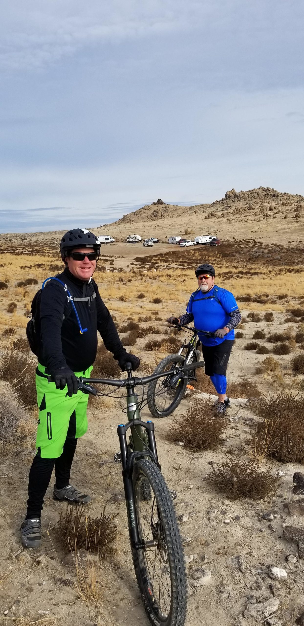 Two men are standing on a dirt trail in a rocky, arid landscape. One man, wearing a black long-sleeve shirt, helmet, and vibrant green shorts, is smiling and leaning on his mountain bike. The other man, dressed in a blue shirt and black shorts, is also smiling while holding his bike. In the background, there are sparse bushes, and several parked vehicles are visible on a distant road under a cloudy sky. Wilson Creek Area mountain bike trail.