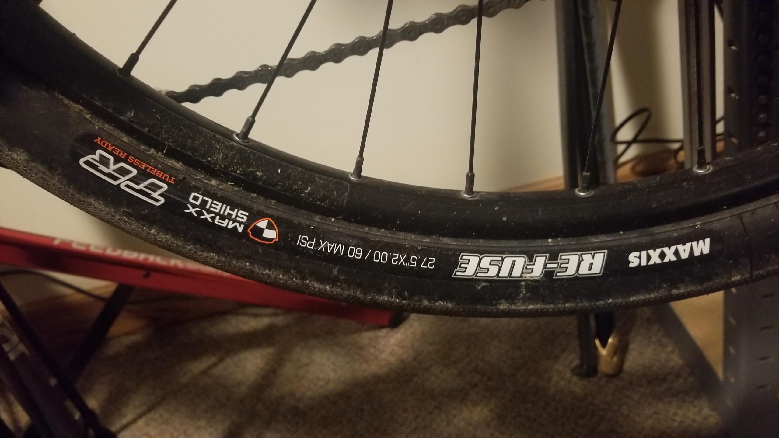 Maxxis Re-Fuse: Close-up of a bicycle tire mounted on a wheel, showing the tread pattern and specifications. The tire is labeled "Maxxis" with the model "RE-FUSE," indicating it is tubeless ready. The image also displays size information: "27.5 x 2.25" and a maximum pressure of 60 PSI. The background features part of a bike stand and flooring.