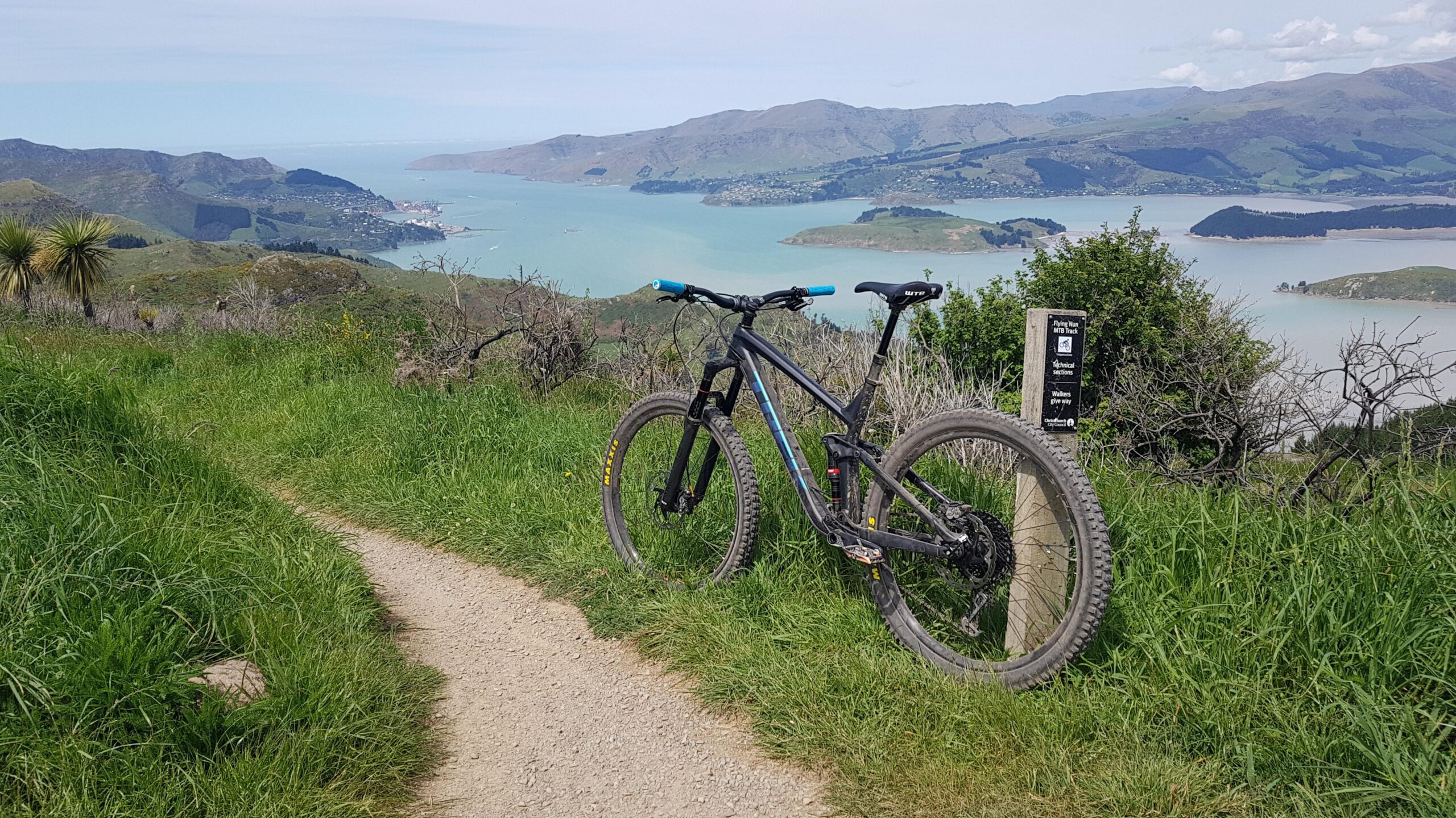 Trek Slash 8: A mountain bike parked beside a dirt path, surrounded by lush green grass and hills. In the background, a scenic view of a bay with blue water and distant mountains. A signpost nearby provides information about the area.