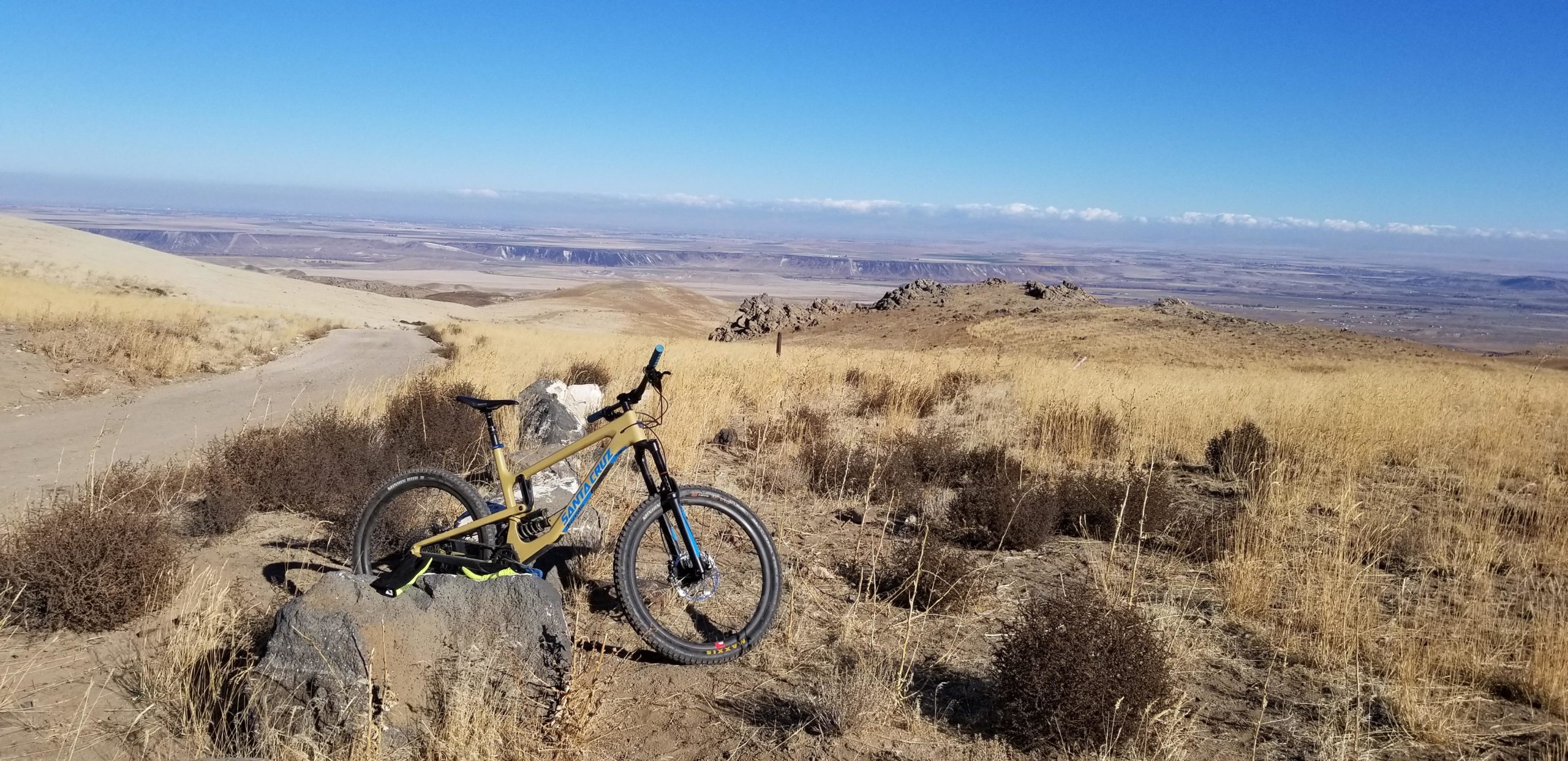 A mountain bike resting on a large rock, surrounded by dry grass and shrubs on a hillside. In the background, a vast landscape extends into the distance with rolling hills and a clear blue sky. Dusty trails lead off into the horizon. Wilson Creek Area mountain bike trail.