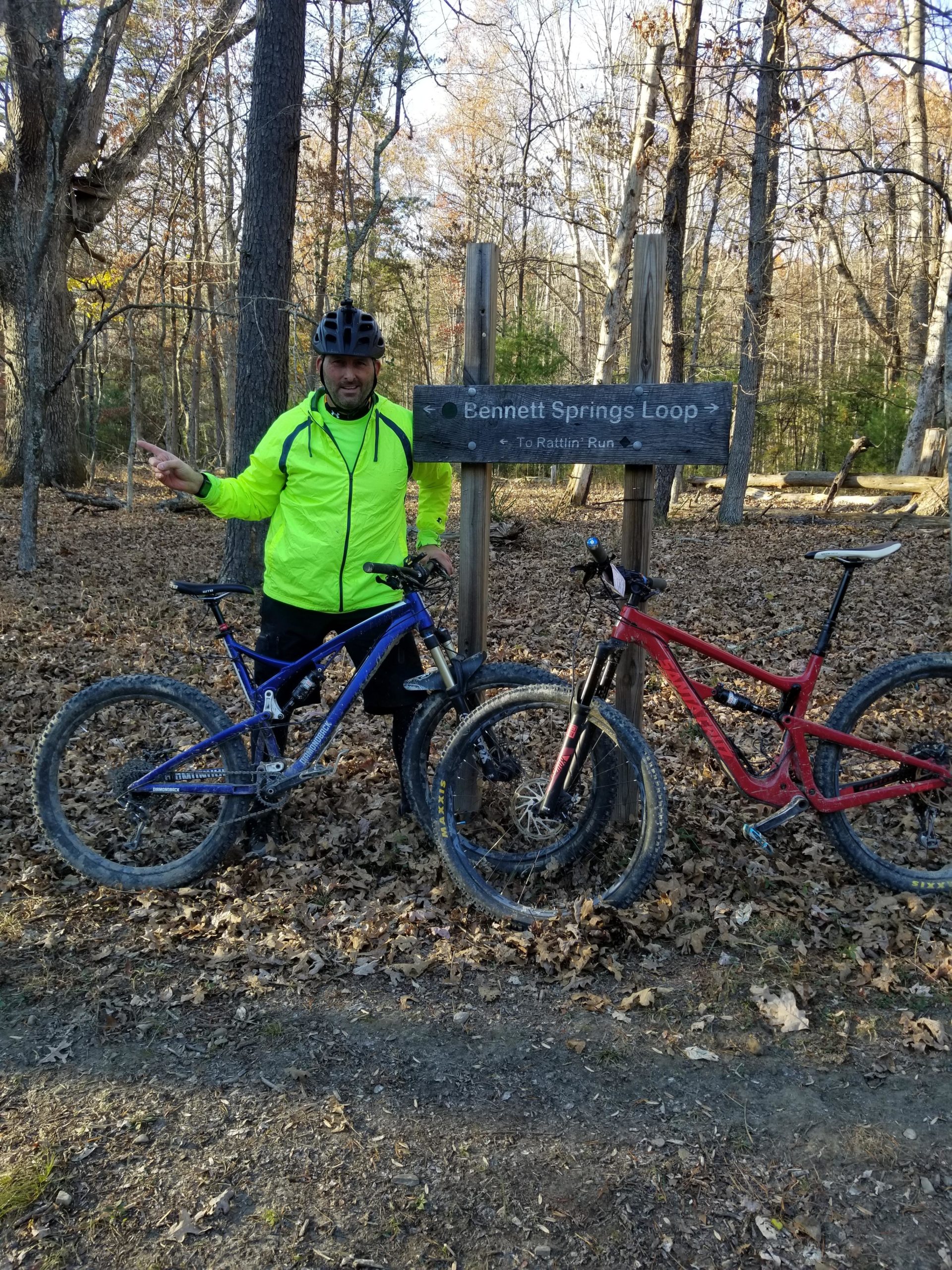 A person wearing a bright yellow jacket and a black helmet stands next to two mountain bikes, one blue and one red, at a trail sign that reads "Bennett Springs Loop." The background features a wooded area with autumn leaves on the ground. Carvin's Cove Trail system mountain bike trail.