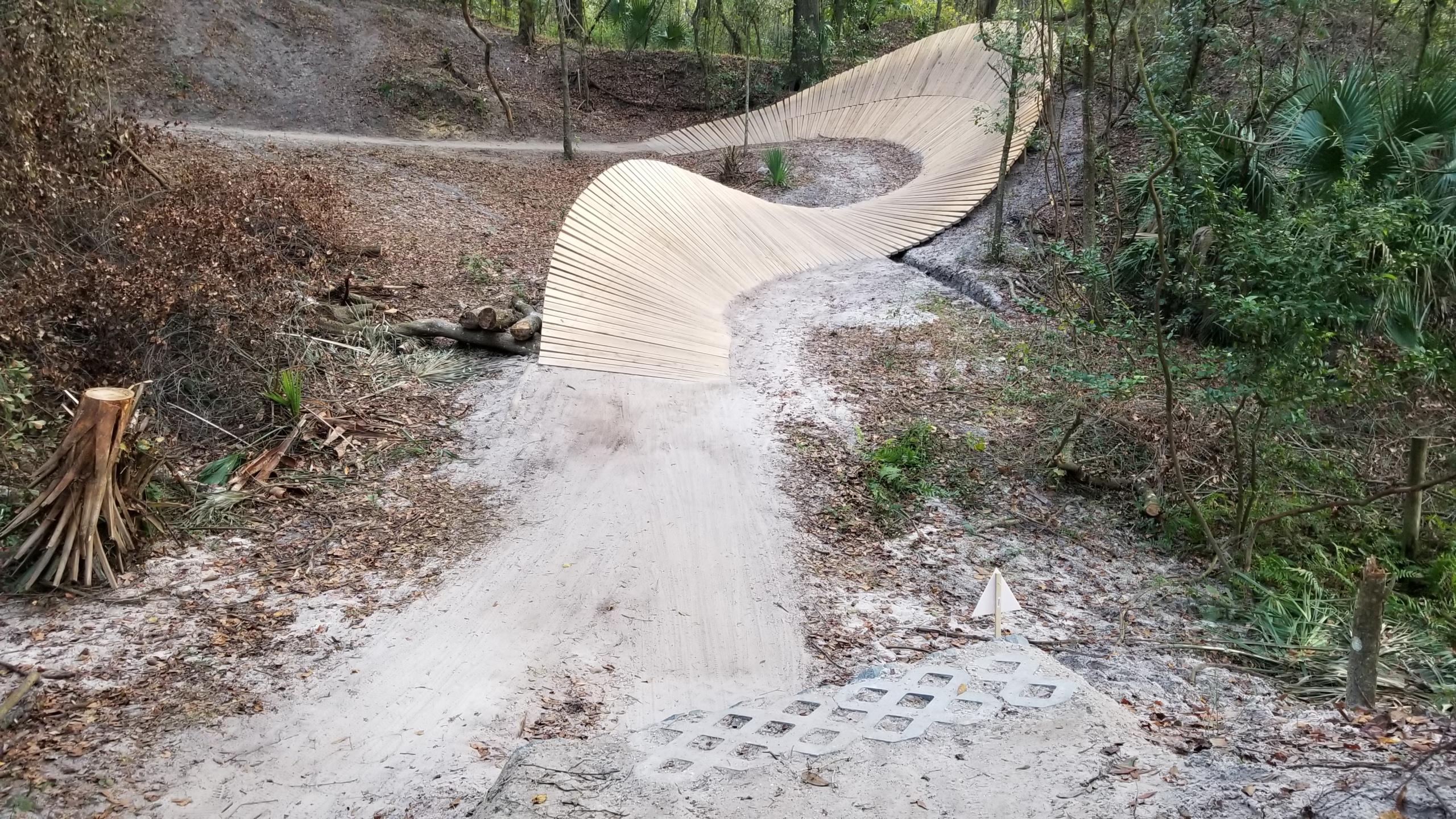 A winding wooden path curves through a forested area, with sandy trails and patches of dry leaves visible on either side. The path features a smooth surface and leads over a natural creek with fallen logs in the foreground. Lush greenery surrounds the trail, creating a peaceful, outdoor setting. Alafia River State Park mountain bike trail.