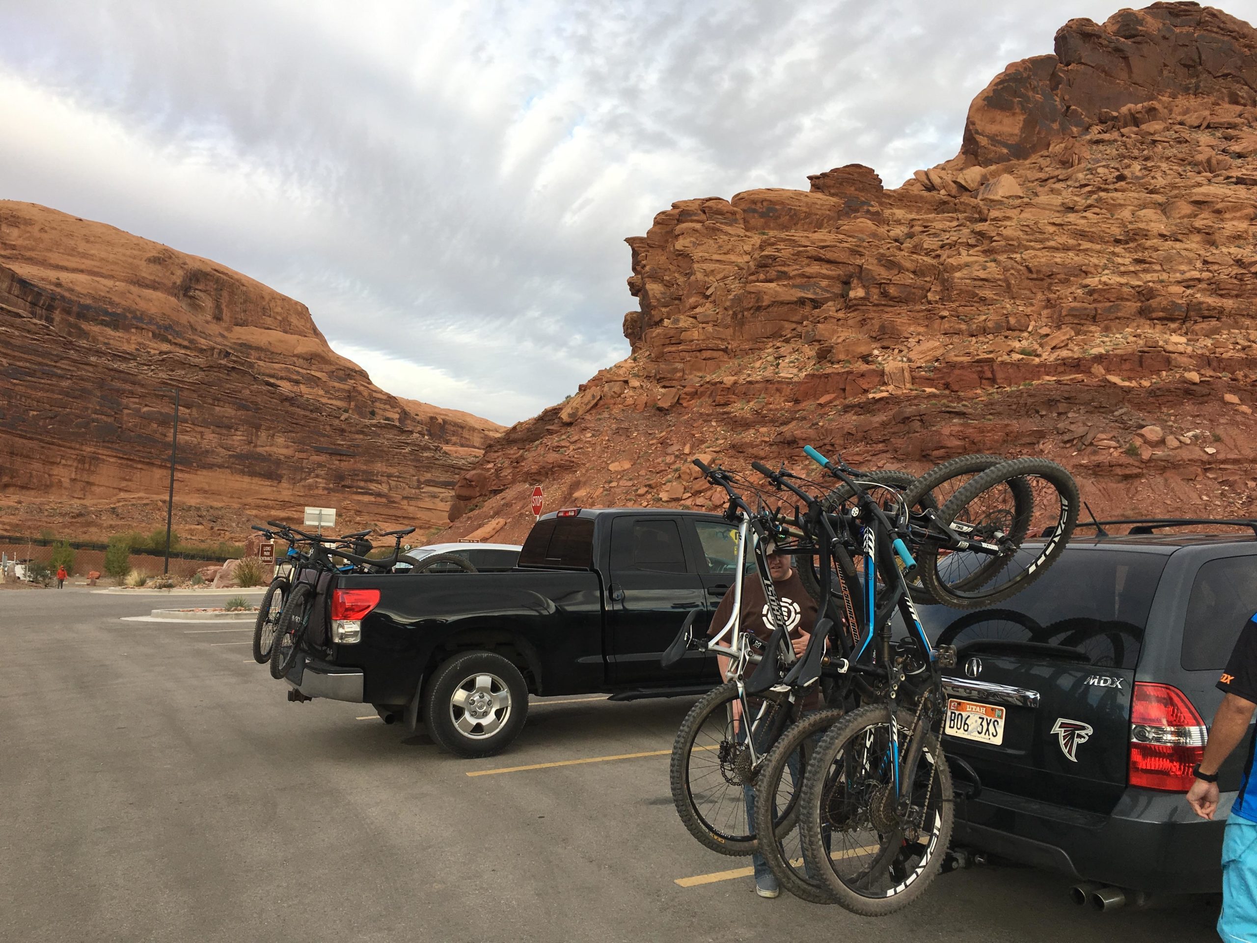 A parking area with two vehicles parked side by side, one a black pickup truck and the other a black SUV, both equipped with mountain bikes mounted on racks. The background features striking red rock formations under a partially cloudy sky, indicating a scenic outdoor location. Porcupine Rim mountain bike trail.