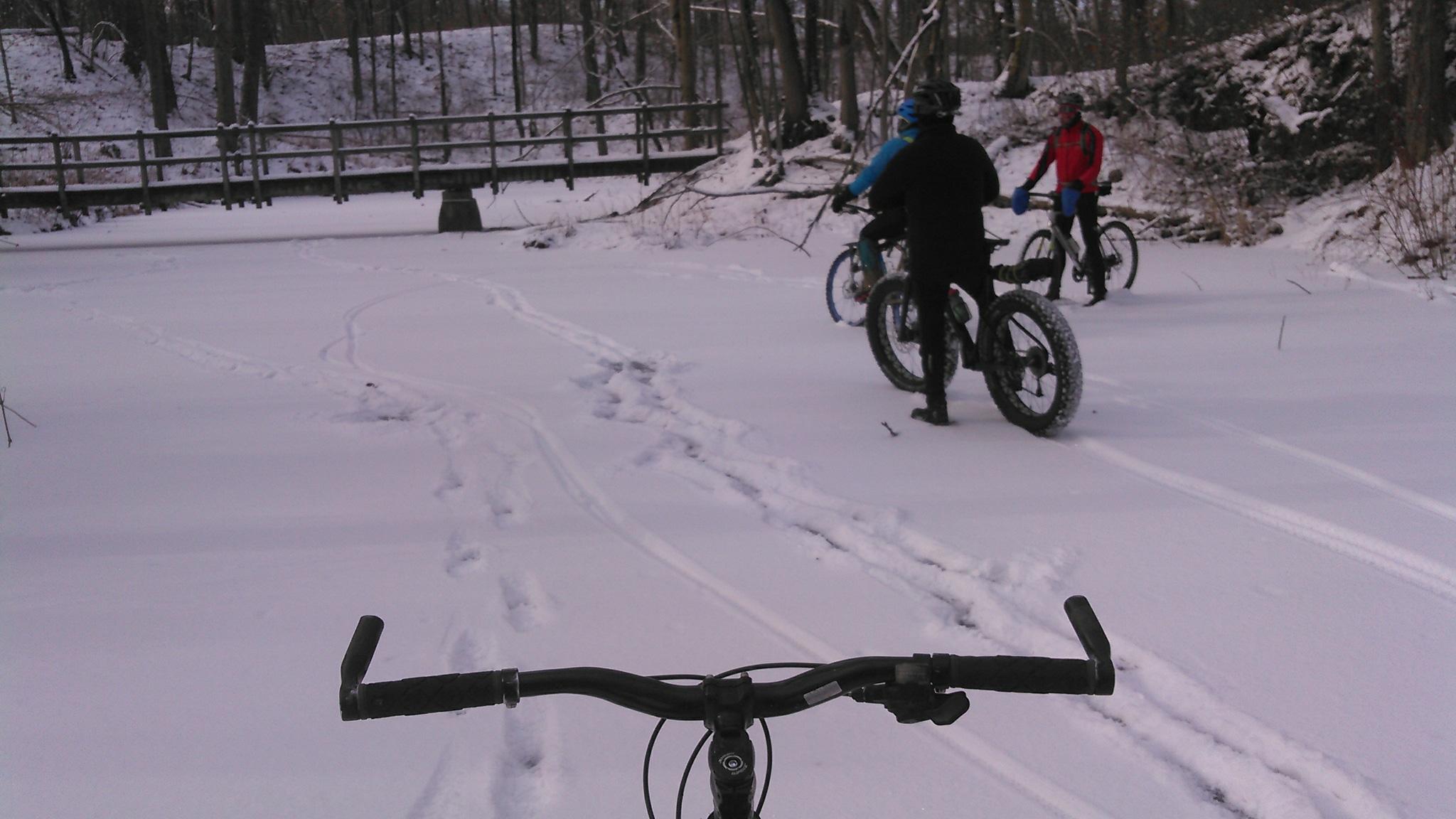 A snowy landscape with bicycle tracks in the fresh snow. The handlebars of a bike are in the foreground, while several cyclists are seen in the background, some adjusting their gear. A wooden bridge can be seen in the distance, surrounded by trees. Moraine State Park mountain bike trail.