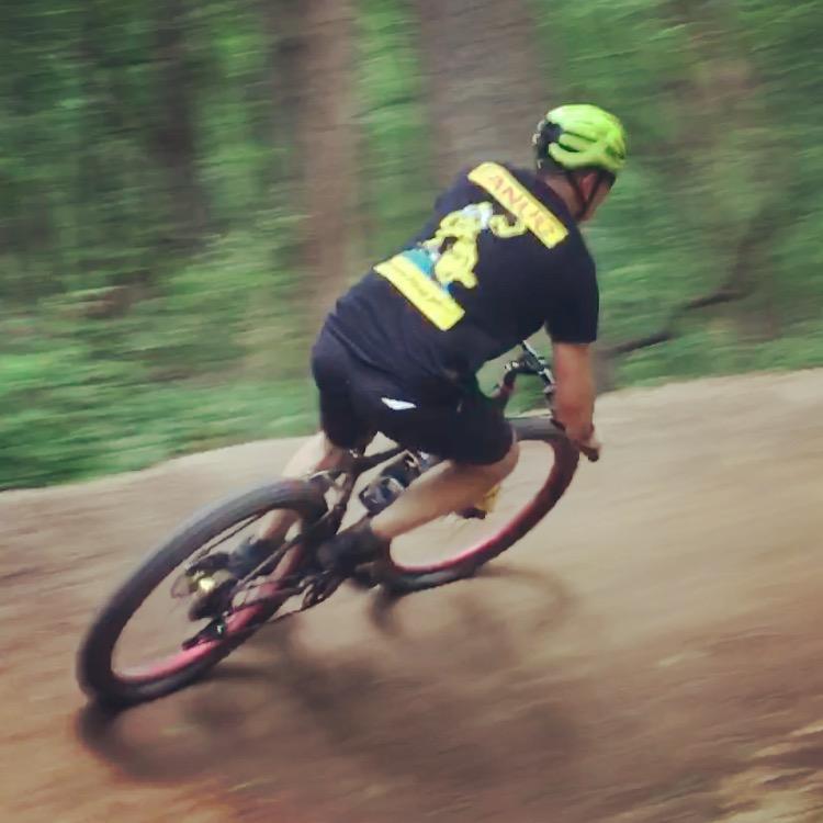 A person riding a mountain bike on a dirt trail, leaning into a turn. The cyclist wears a bright green helmet and a black t-shirt with graphics on the back. The background features lush green trees, indicating a forested area. The motion blur suggests speed and excitement. DTE Energy Foundation Trail mountain bike trail.