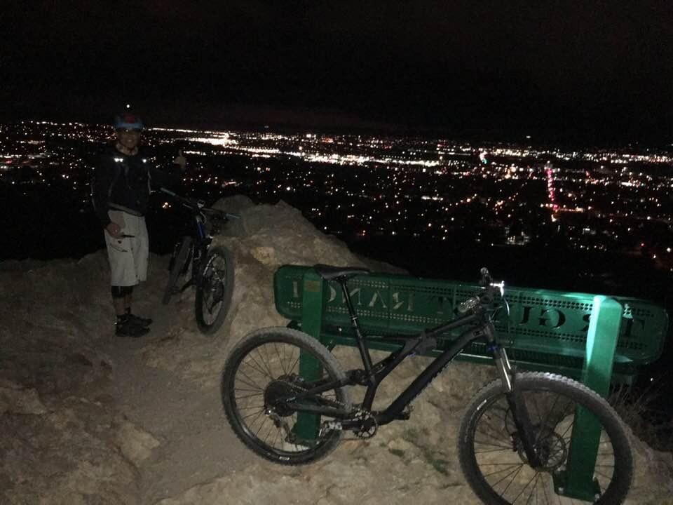 A person standing next to two mountain bikes on a rocky outcrop at night, overlooking a city illuminated by lights. The individual is wearing a helmet and gear, giving a thumbs up. A green bench is visible in the background, featuring a view of the city below. Bonneville Shoreline Trail - Ogden Section mountain bike trail.