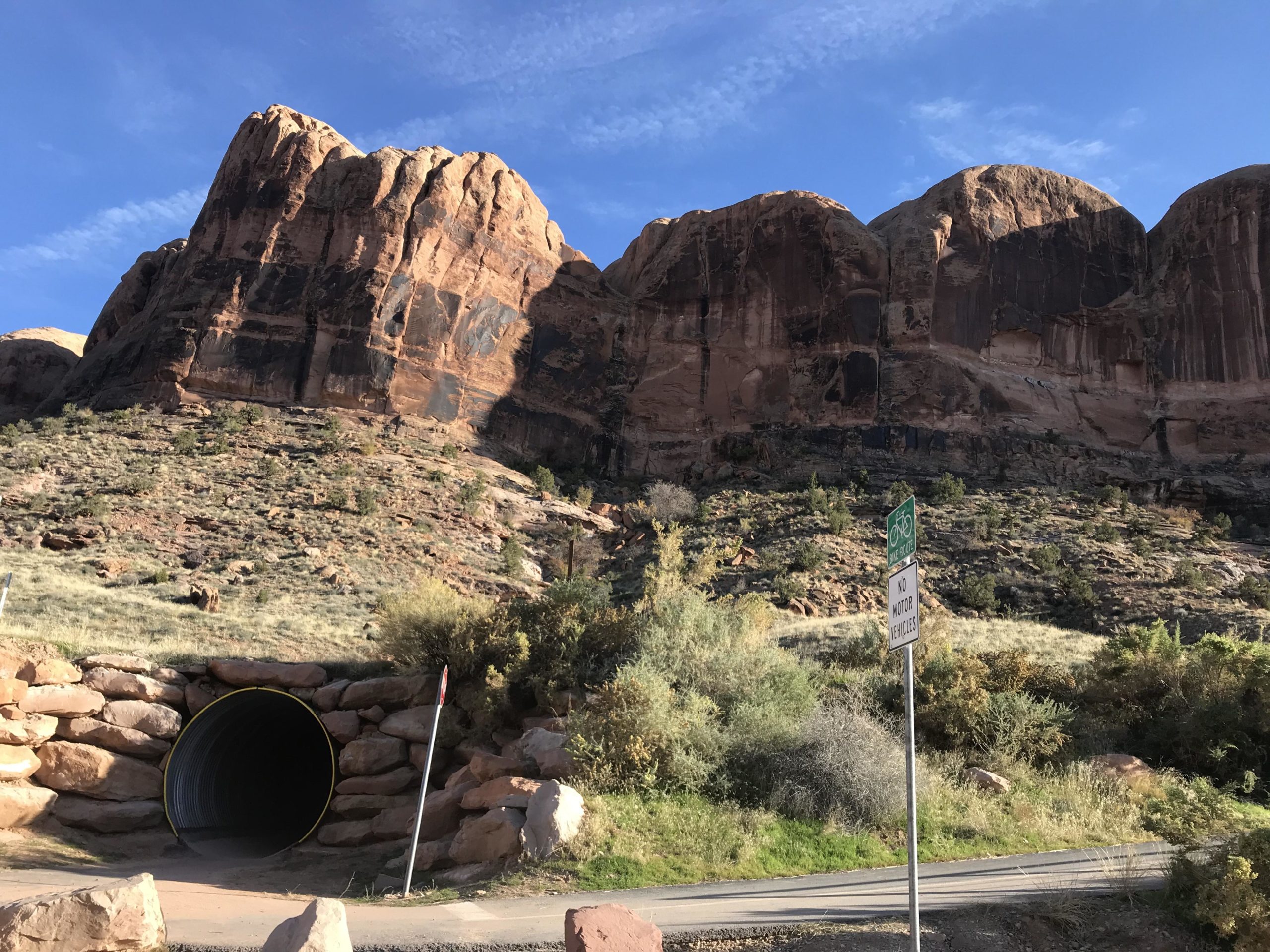 A scenic view featuring rugged rock formations and a metal tunnel entrance at the base, surrounded by greenery and shrubs. The sky above is blue with scattered clouds, while signs indicate a bike path and no motor vehicles. Porcupine Rim mountain bike trail.