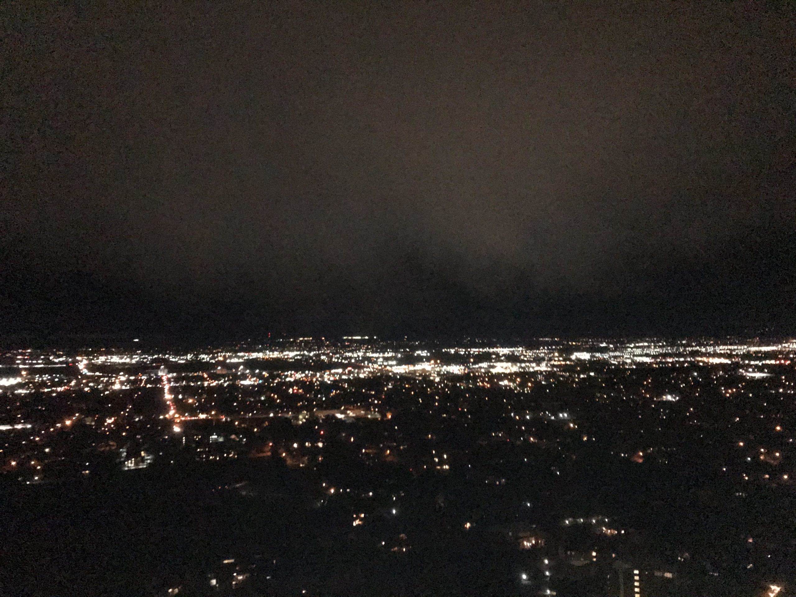 A night view of a cityscape, showcasing a wide expanse of twinkling lights against a dark sky. The scene captures illuminated streets and buildings, with a backdrop of clouds partially obscuring the stars. Bonneville Shoreline Trail - Ogden Section mountain bike trail.