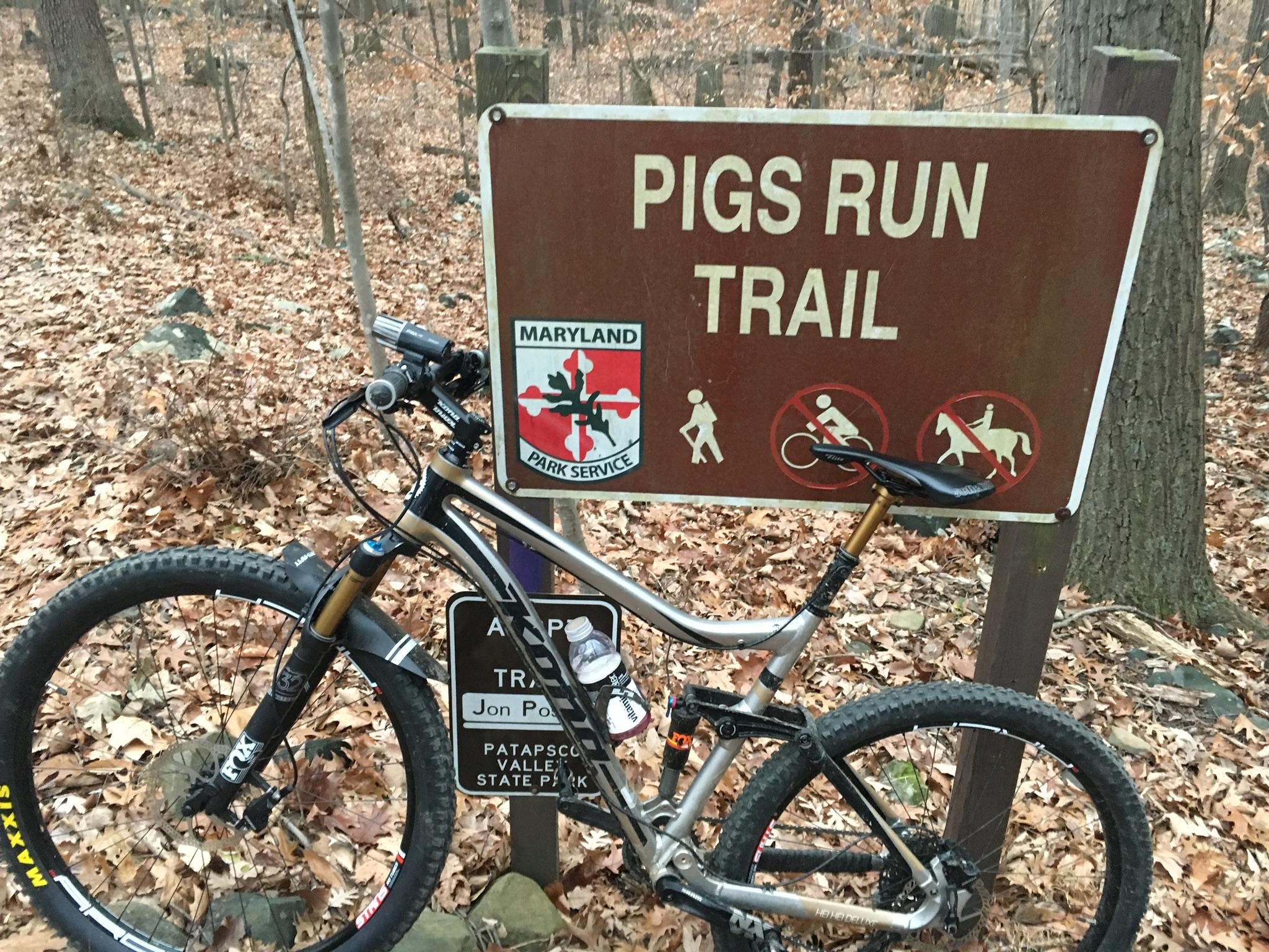 A mountain bike parked next to a trail sign that reads "Pigs Run Trail." The sign features the Maryland Park Service logo and includes icons indicating that's the trail is open for hiking but not for biking or horseback riding. The surrounding area is wooded with fallen leaves covering the ground. Patapsco Valley State Park (Avalon Area) mountain bike trail.