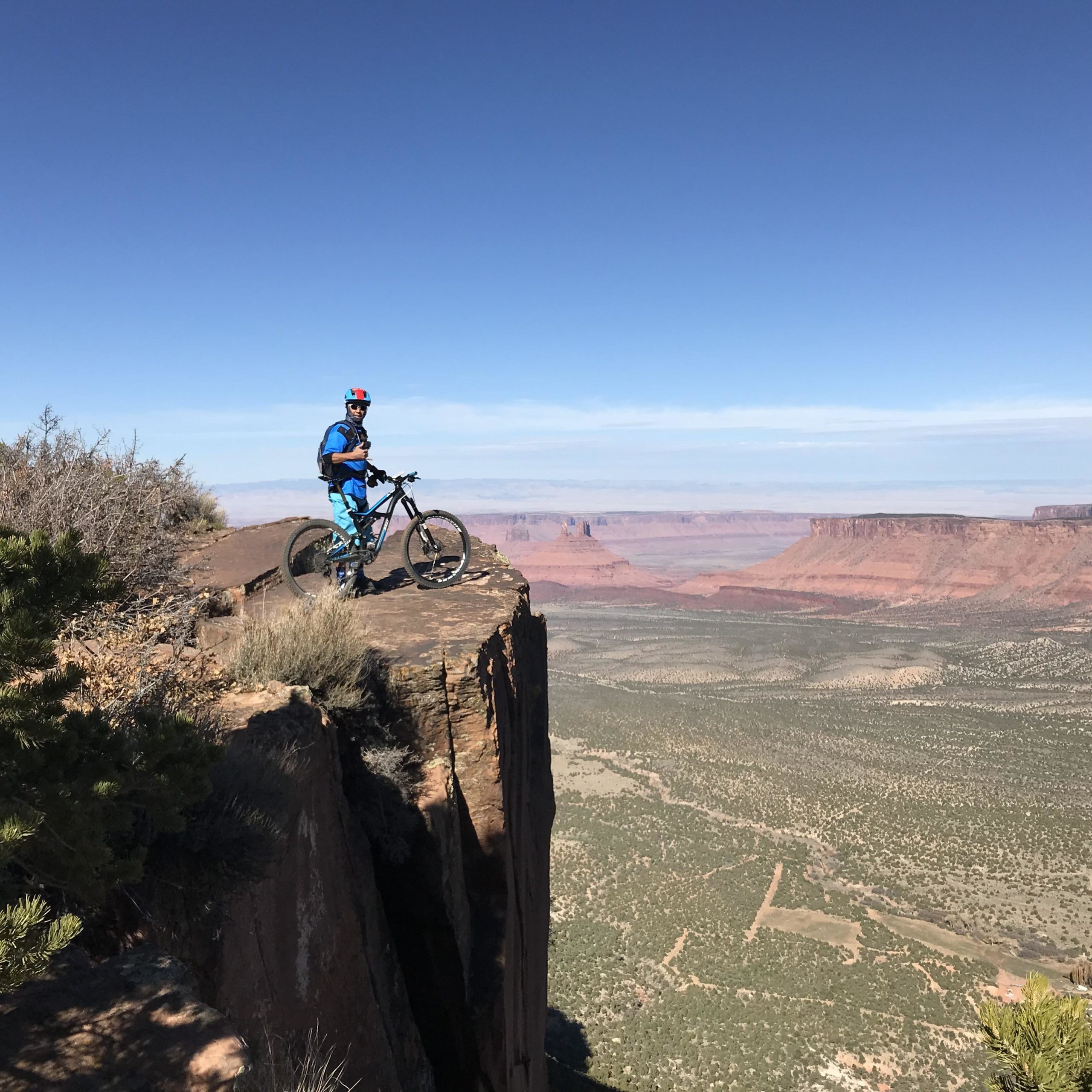 A mountain biker standing on a rocky ledge with a panoramic view of red rock formations and desert landscape in the background under a clear blue sky. Porcupine Rim mountain bike trail.