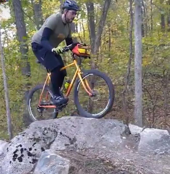 A person riding a yellow mountain bike, wearing a helmet and gloves, is jumping over a large rock in a wooded trail surrounded by trees with autumn foliage. DTE Energy Foundation Trail mountain bike trail.