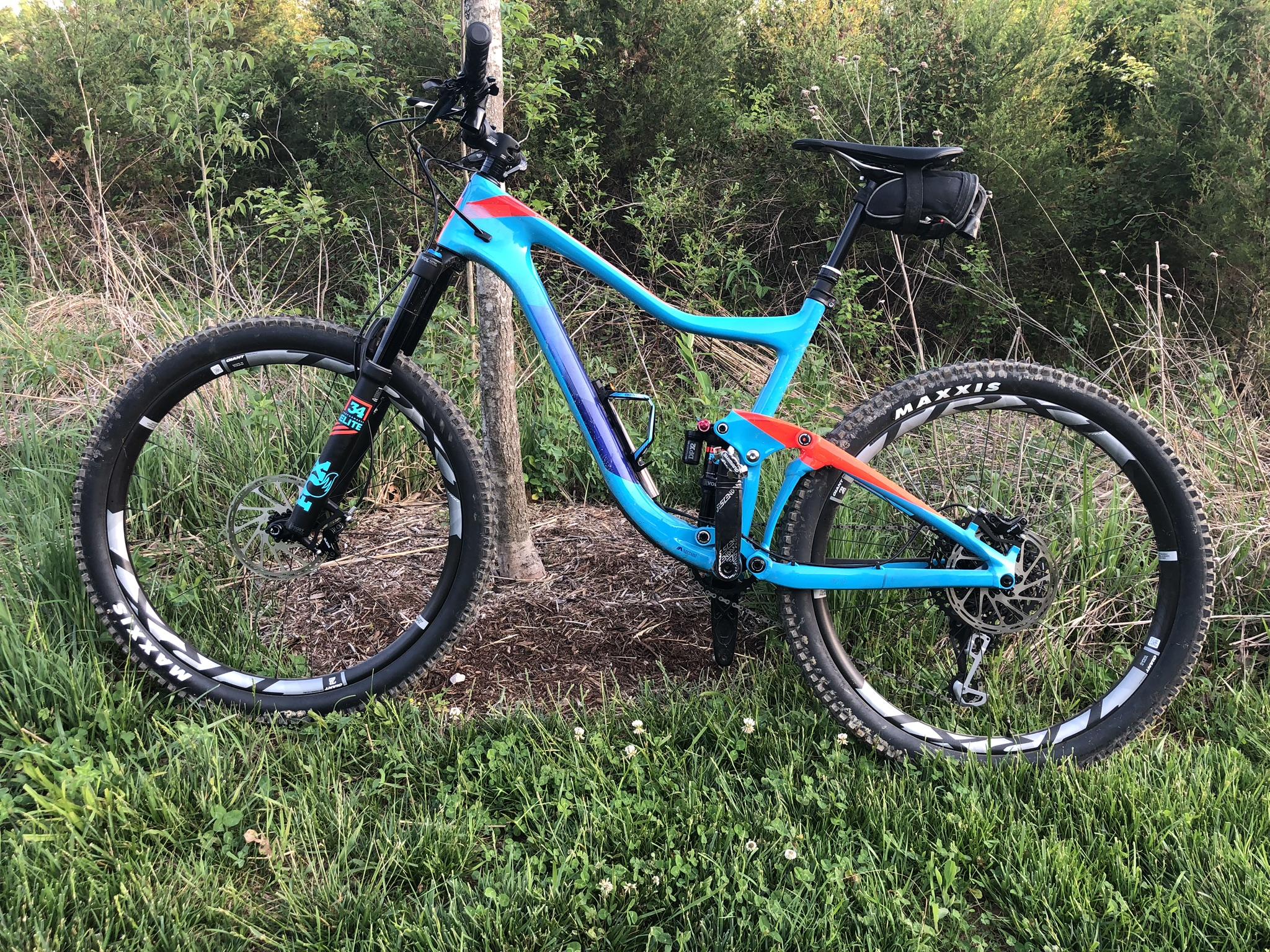 Giant Giant Trance Advanced 27.5 2: A vibrant blue mountain bike with red accents leans against a tree in a grassy area, surrounded by greenery. The bike features thick tires, a front suspension fork, and a black seat bag, indicating that it is designed for off-road trails and rugged terrains.