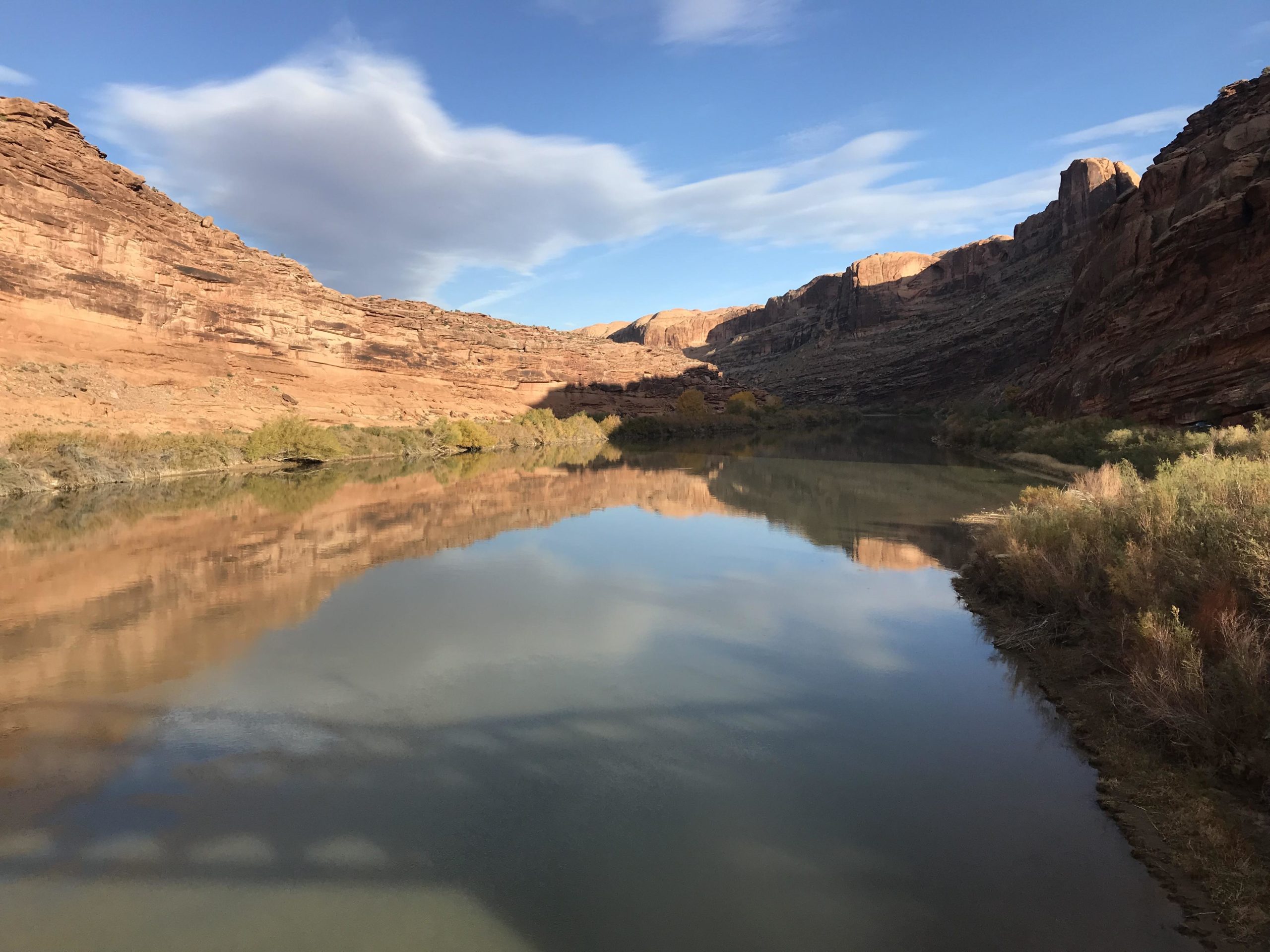 A tranquil river scene surrounded by towering red rock cliffs under a blue sky with scattered clouds. The calm water reflects the cliffs and nearby vegetation, creating a serene natural landscape. Porcupine Rim mountain bike trail.