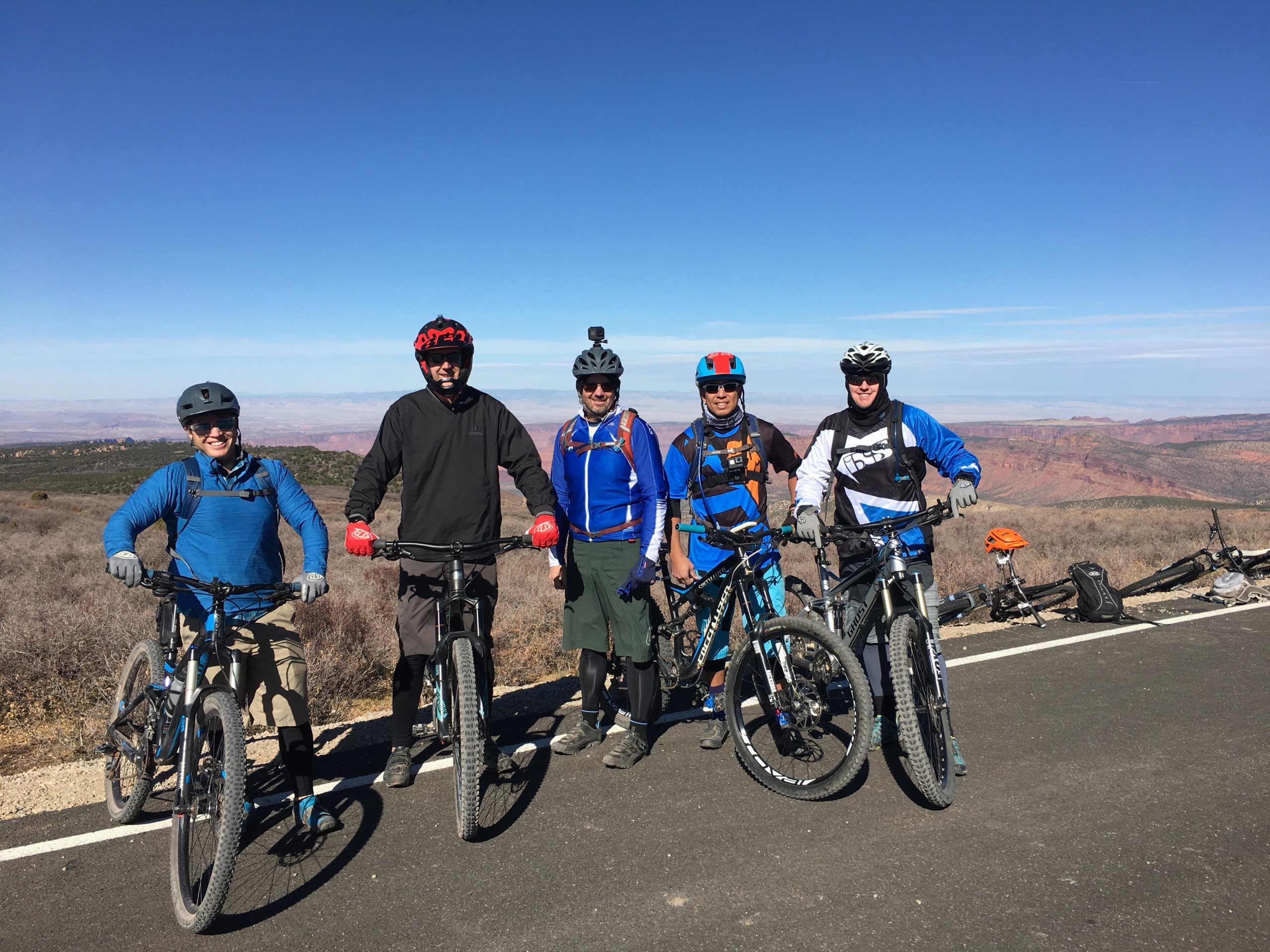 Five mountain bikers pose for a photo on a scenic overlook with a vast landscape in the background. They are wearing helmets and biking gear, standing beside their bicycles on a sunny day. The terrain features shrubs and rocky formations, and distant hills are visible under a clear blue sky. Porcupine Rim mountain bike trail.