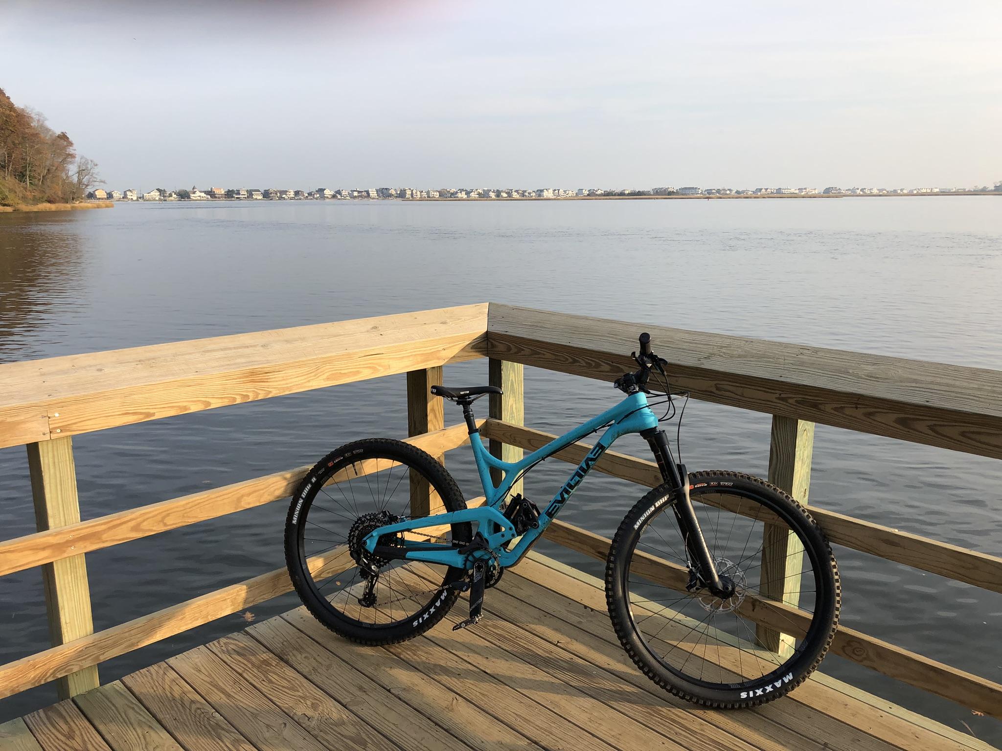 A mountain bike rests against the railing of a wooden pier, overlooking a calm body of water. The scene features a tranquil view with houses visible in the distance across the water, surrounded by trees and a clear sky. Hartshorne Woods Park mountain bike trail.