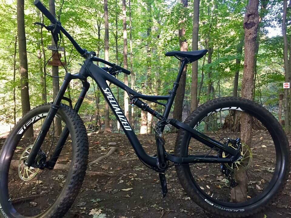 Specialized Stumpjumper FSR: A black mountain bike with wide tires parked next to a tree in a forested area, featuring lush green foliage in the background. The bike has a sleek design and displays the logo "Specialized" prominently on its frame.