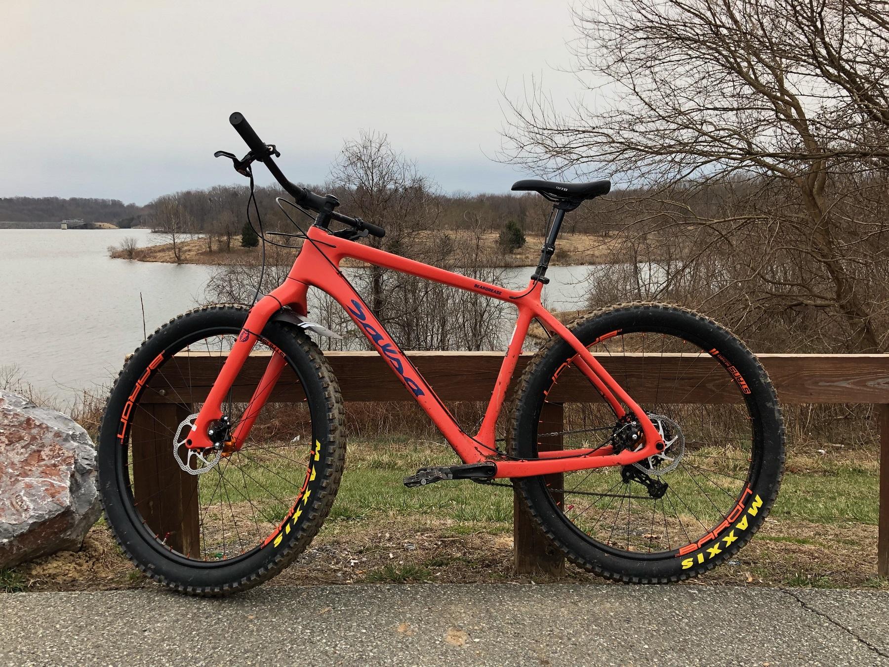 Salsa Beargrease: A bright orange mountain bike is parked alongside a wooden railing near a calm lake. The bike features chunky tires with visible branding and has a scenic backdrop of trees and a cloudy sky.