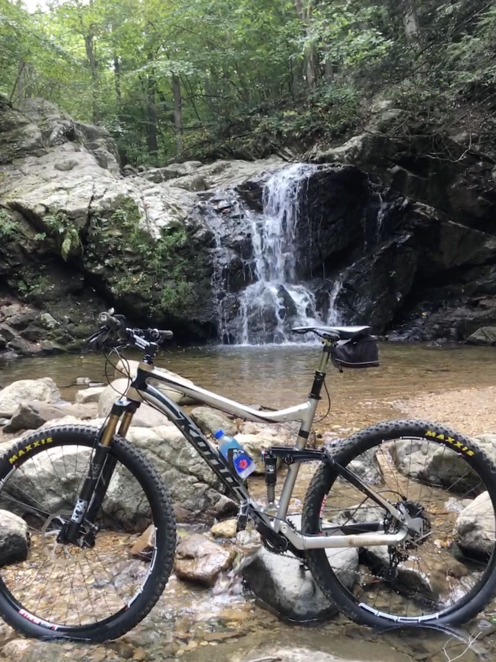 A mountain bike parked on a rocky riverbank near a small waterfall, surrounded by lush green trees and natural scenery. The bike is positioned on stones near the water, with a water bottle attached to its frame. Patapsco Valley State Park (Avalon Area) mountain bike trail.
