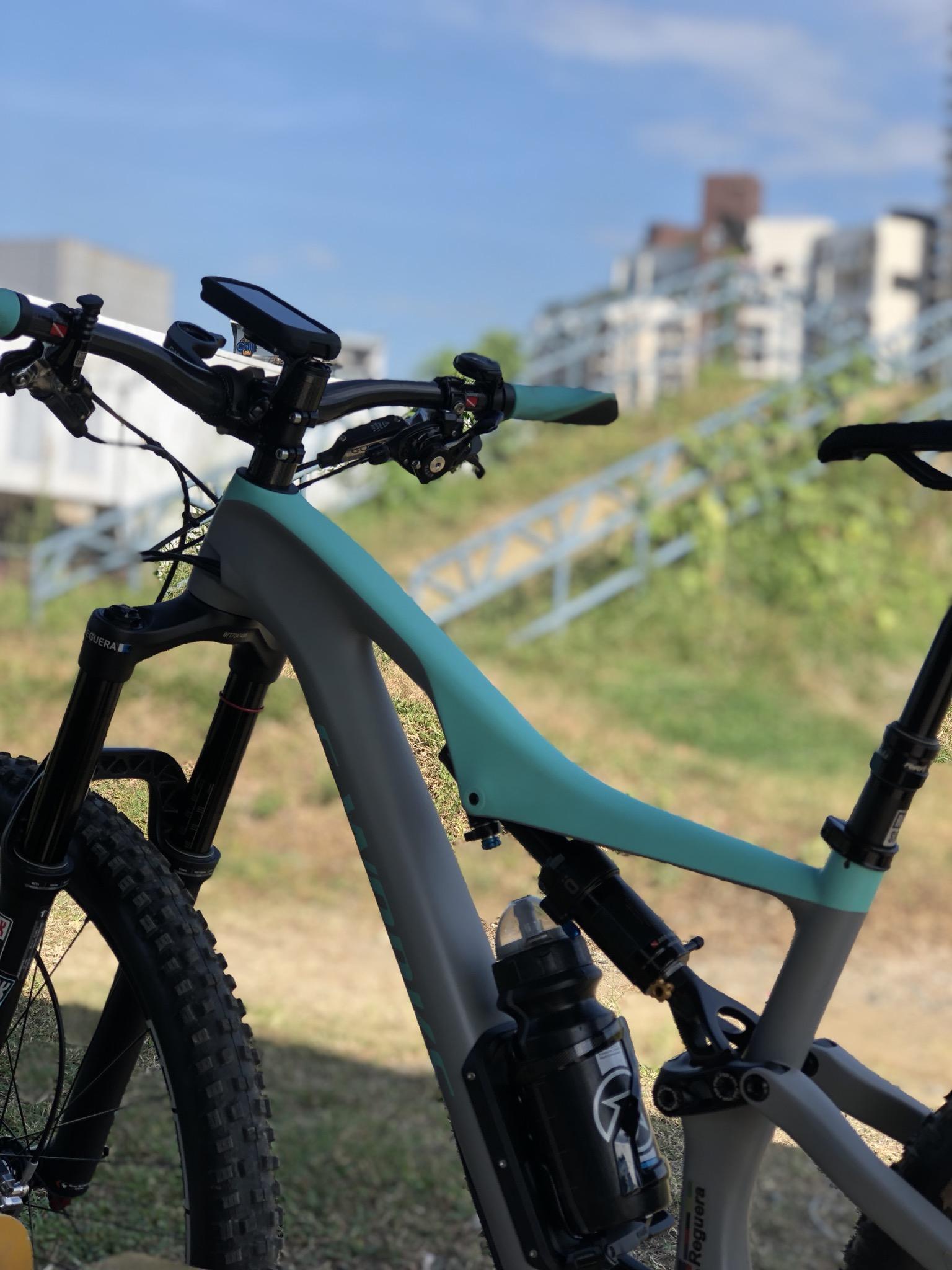 Specialized Stumpjumper: A close-up view of a mountain bike's handlebars and frame, featuring a light blue accent on the gray frame. The bike is positioned outdoors with a grassy area in the background. A water bottle is mounted on the frame, and a staircase can be seen in the distance. The sky is clear with a few clouds.