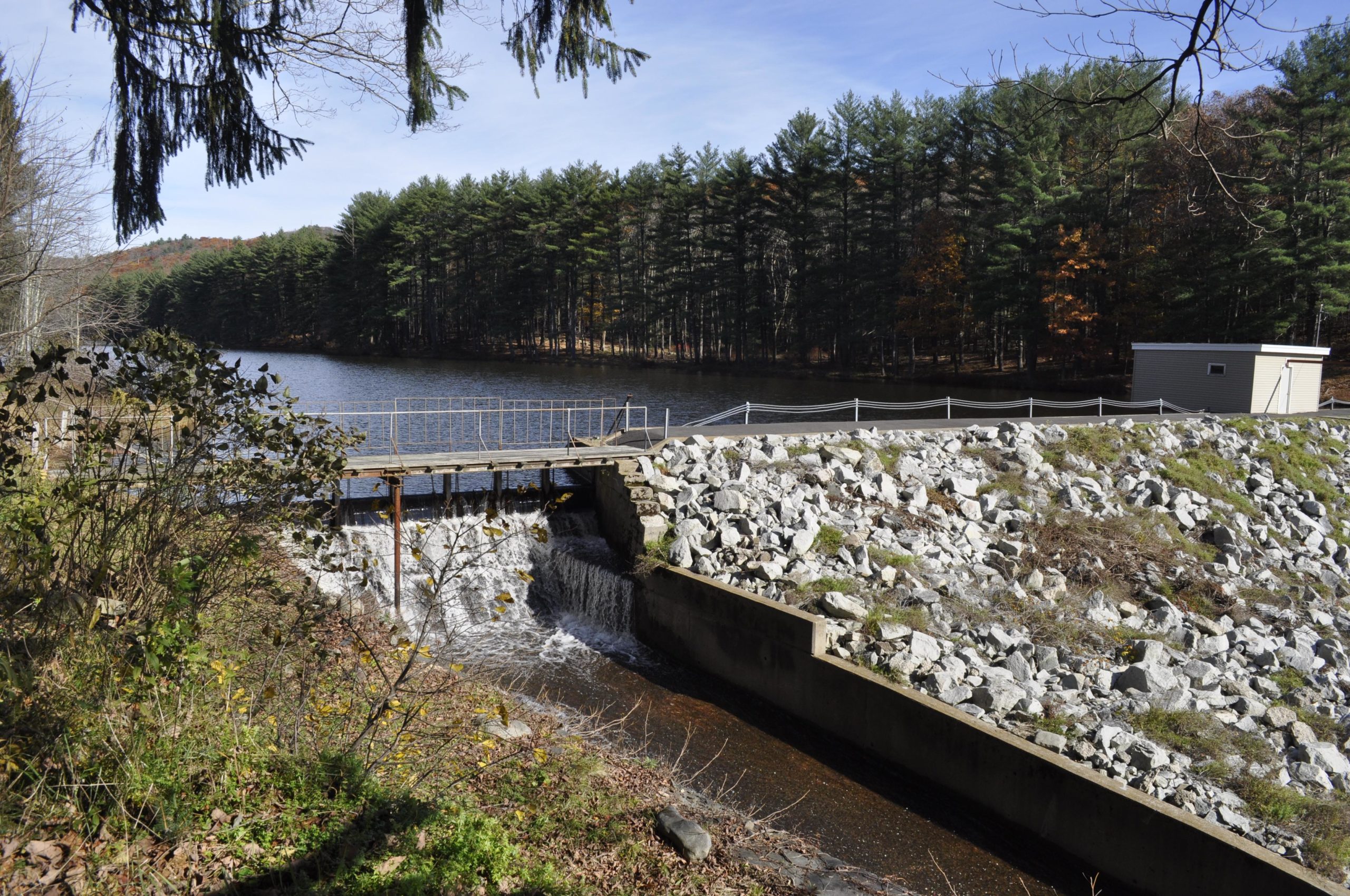 A serene landscape featuring a small dam with water flowing over it, surrounded by rocky terrain and lush greenery. The dam is situated next to a calm body of water, with a backdrop of pine trees and a clear blue sky. A small building is visible near the dam, contributing to the natural scene. Watershed Park mountain bike trail.