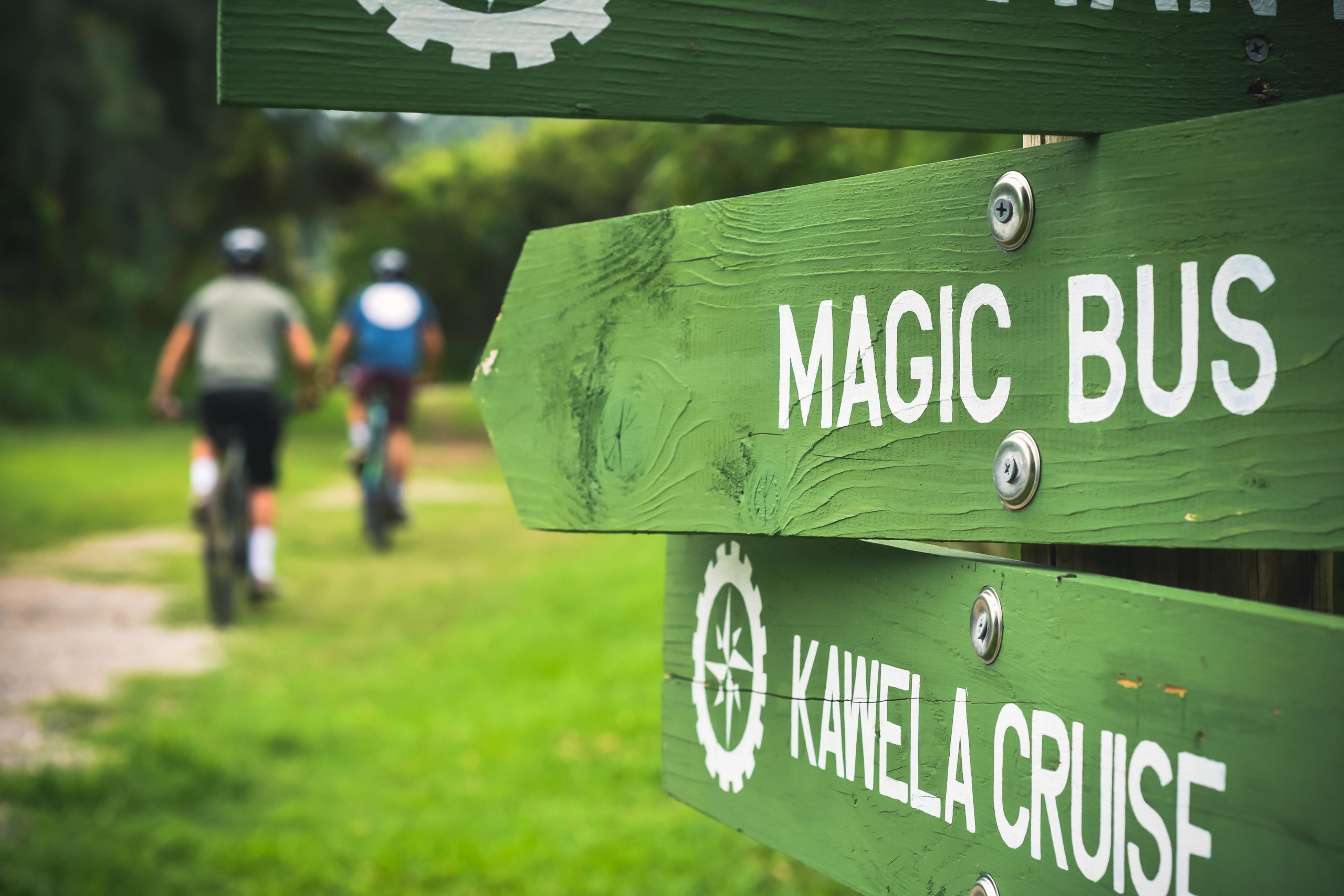 A green directional sign pointing to "Magic Bus" and "Kawela Cruise," with two cyclists in the background riding on a grassy path. Magic Bus Loop mountain bike trail.