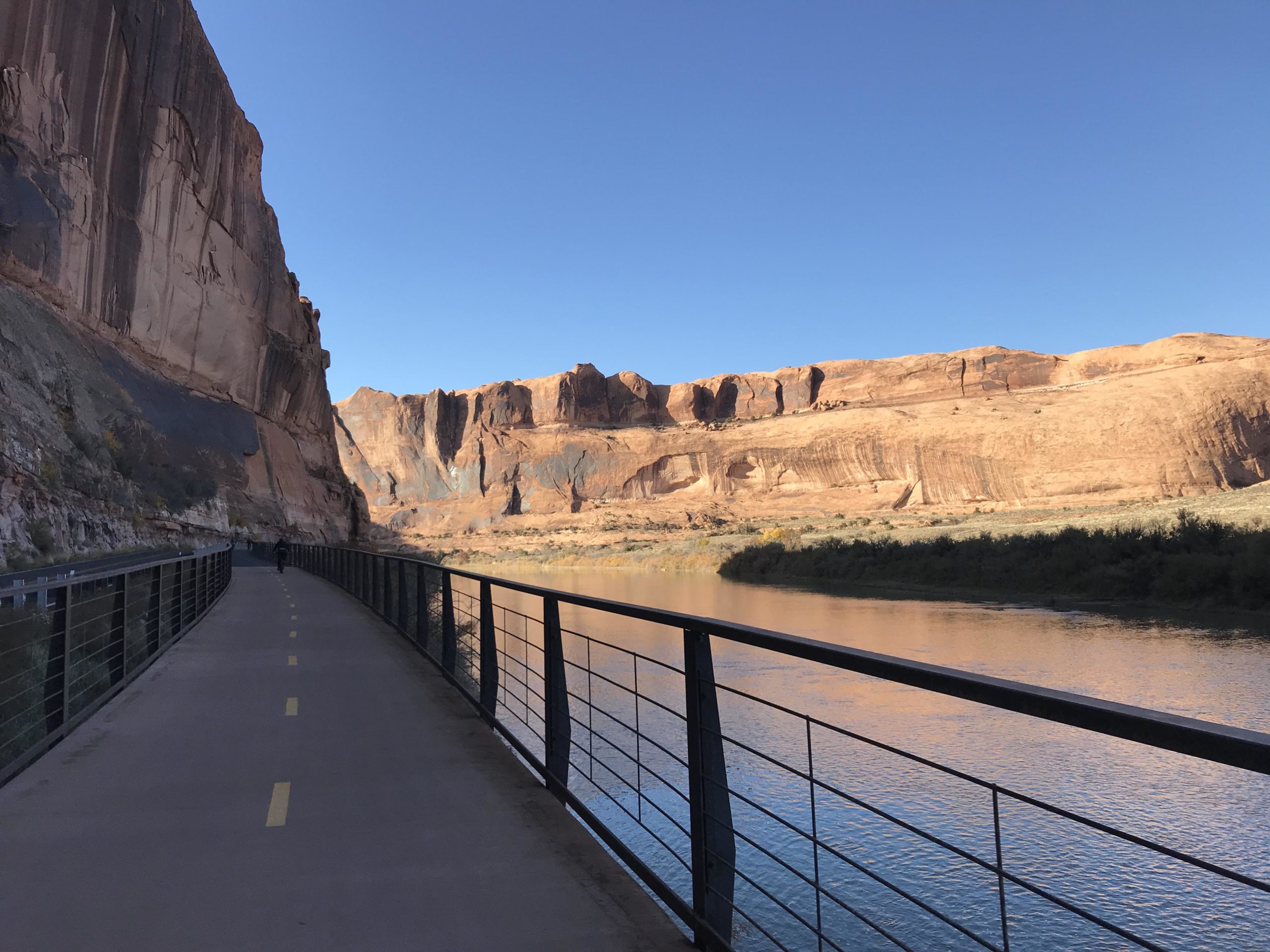 A scenic view of a paved path alongside a river, bordered by steep cliff formations and rock faces under a clear blue sky. The path includes yellow markings and is lined with a metal railing, while a cyclist can be seen in the distance. The water reflects the colors of the surrounding landscape. Porcupine Rim mountain bike trail.