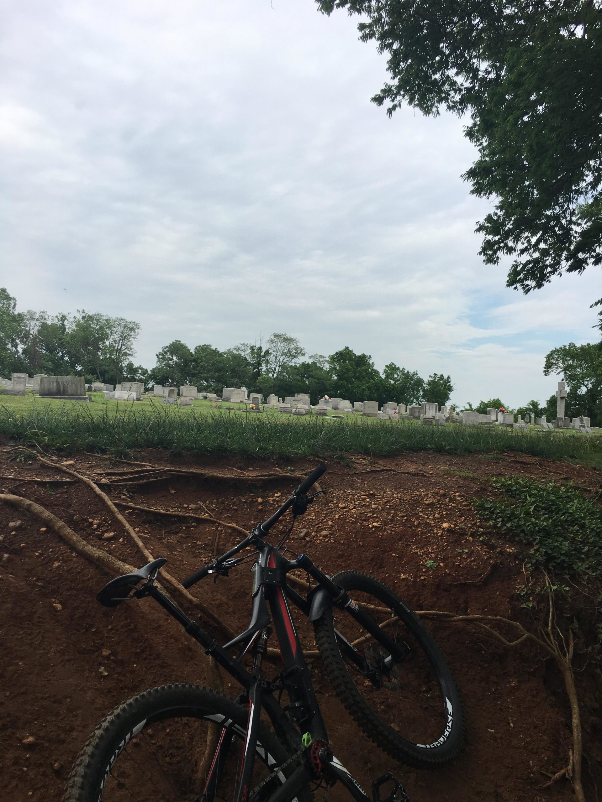 A mountain bike resting against a small dirt embankment, with a cemetery visible in the background under a cloudy sky. Northbank Trail mountain bike trail.