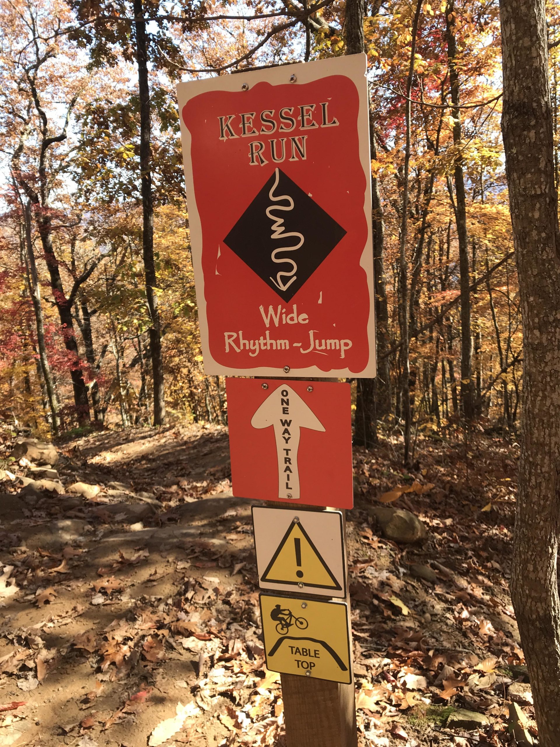 A trail sign in a wooded area displaying information for the "Kessel Run" biking trail, which indicates it features wide rhythm jumps. Additional signs indicate the trail is a one-way route and warn of a tabletop feature, along with a caution symbol. The surrounding trees are adorned with autumn leaves, creating a colorful backdrop. Fire Mountain Trail System mountain bike trail.