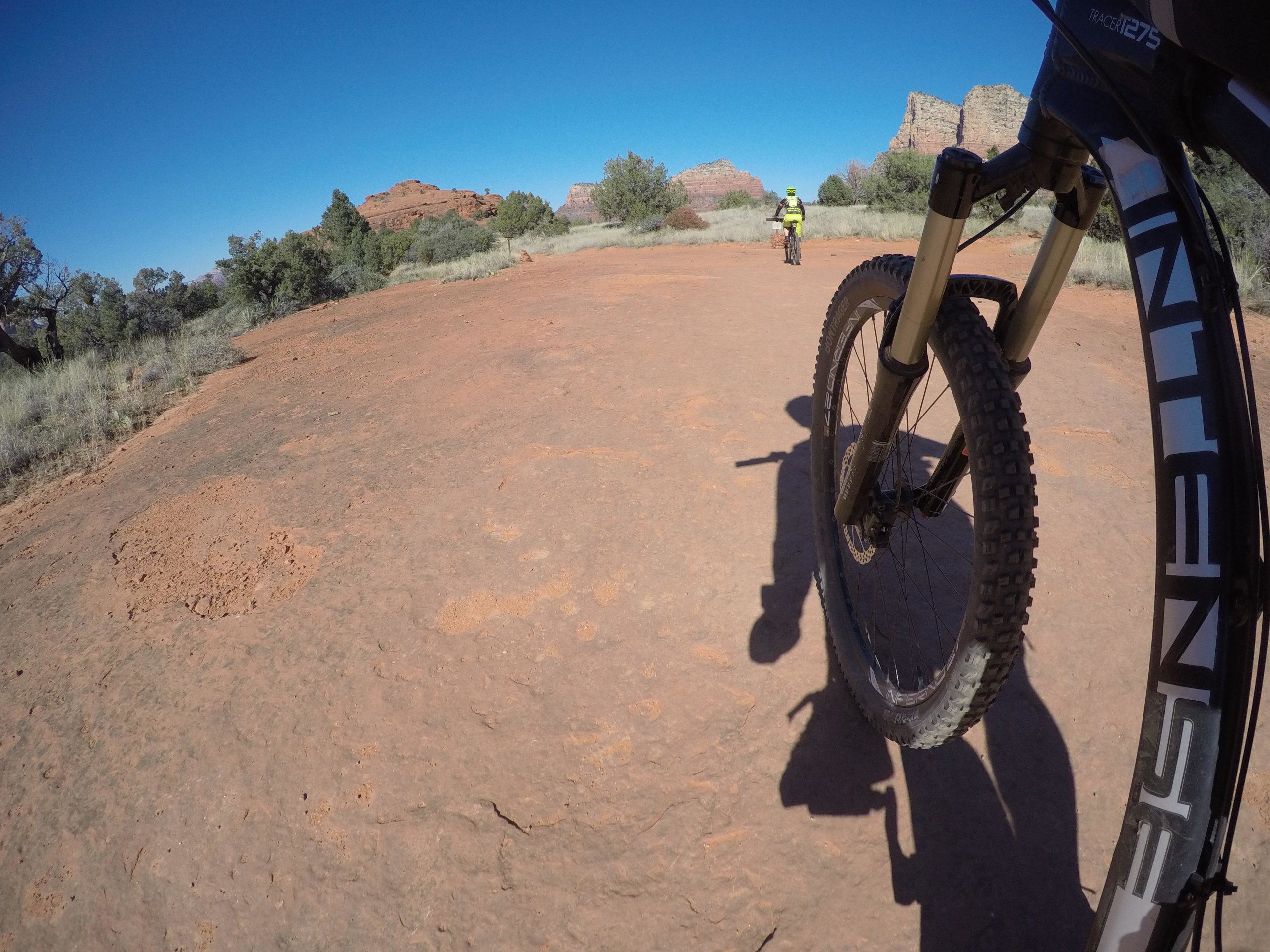 A close-up view of a mountain bike wheel on a rocky trail, with a cyclist in the background riding on a dirt path surrounded by desert vegetation and sandstone formations under a clear blue sky. Bell Rock Area Trails mountain bike trail.