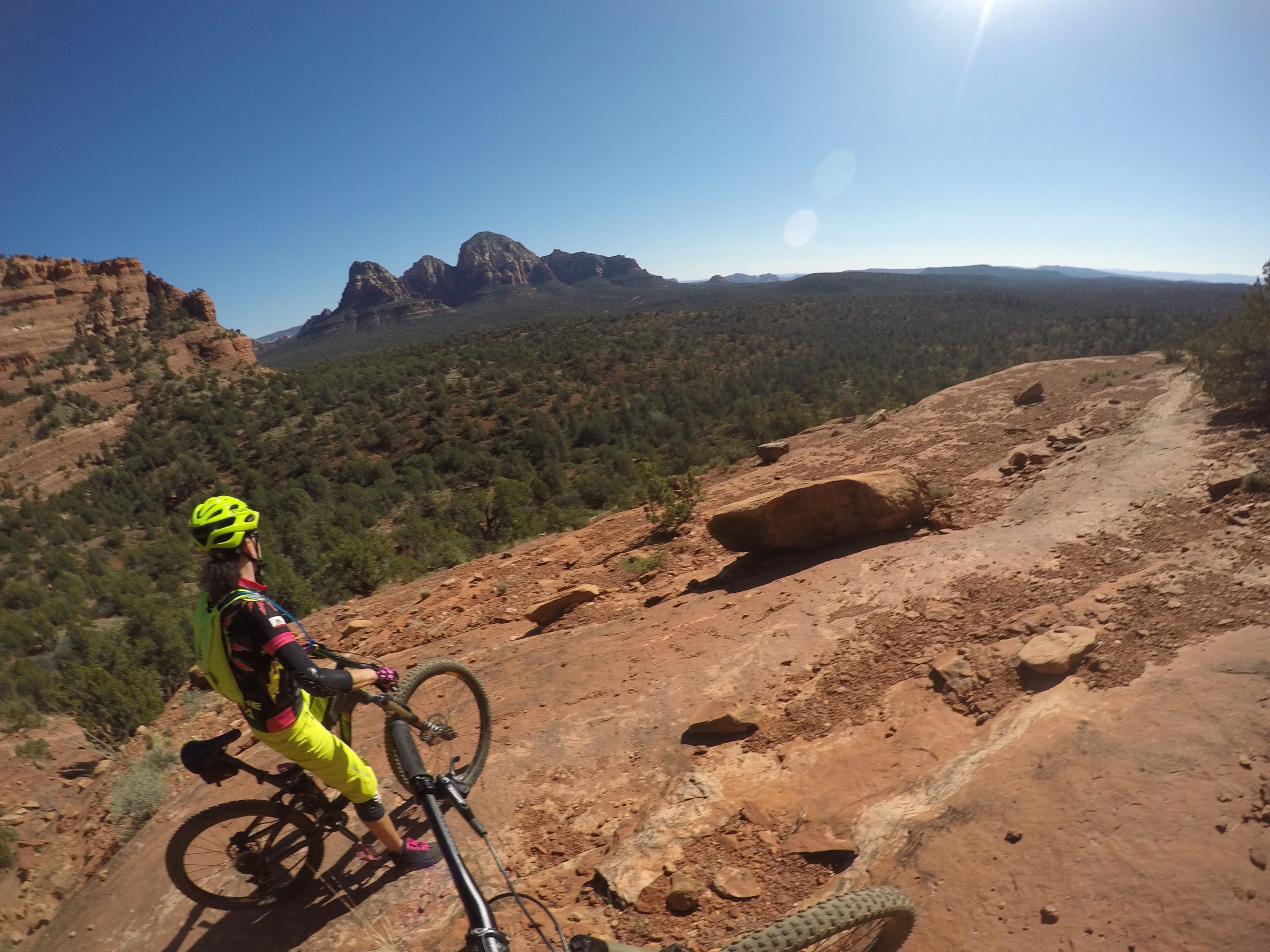 A mountain biker wearing a bright yellow helmet and colorful attire stands on rocky terrain, overlooking a lush forest and distant mountains under a clear blue sky. The biker appears to be assessing the trail ahead. Upper Dry Creek Area Trails mountain bike trail.