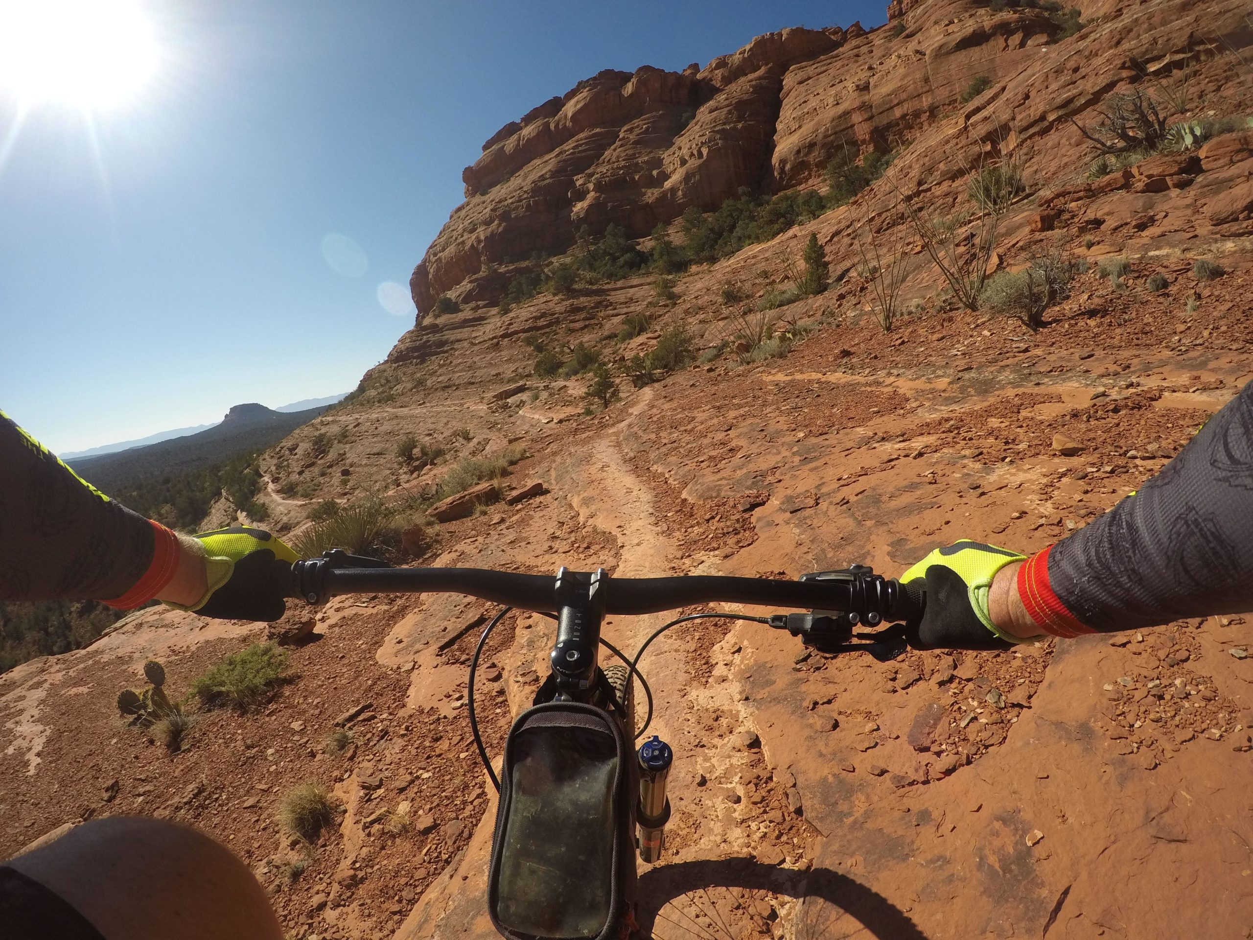 A mountain biker's perspective while riding along a rocky trail in a sunlit desert landscape, with rugged rock formations and sparse vegetation visible in the background. The biker's gloved hands grasp the handlebars, showcasing a close-up view of the bike and the terrain. Upper Dry Creek Area Trails mountain bike trail.