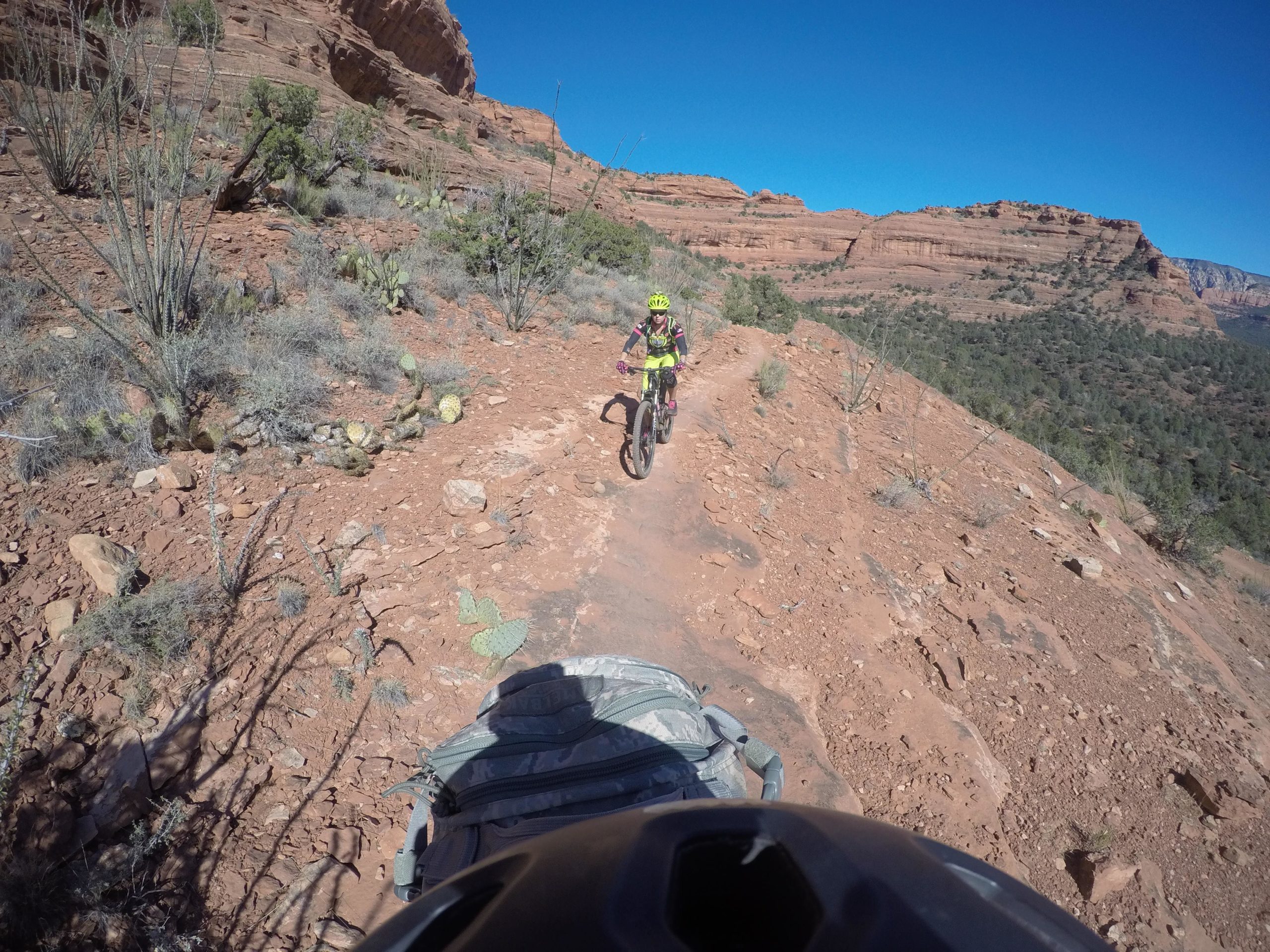 A mountain biker navigating a rocky trail through a desert landscape, with red rock formations and sparse vegetation in the background under a clear blue sky. Upper Dry Creek Area Trails mountain bike trail.