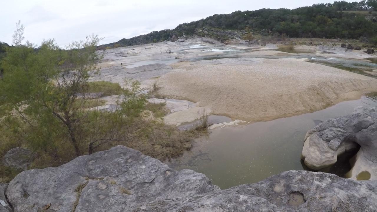 A rocky riverbank scene with sandy areas and shallow water. Sparse vegetation is visible in the foreground, while a group of people can be seen in the distance engaging in outdoor activities. The background features hills and a cloudy sky, suggesting a calm and natural setting. Hackenburg Loop mountain bike trail.