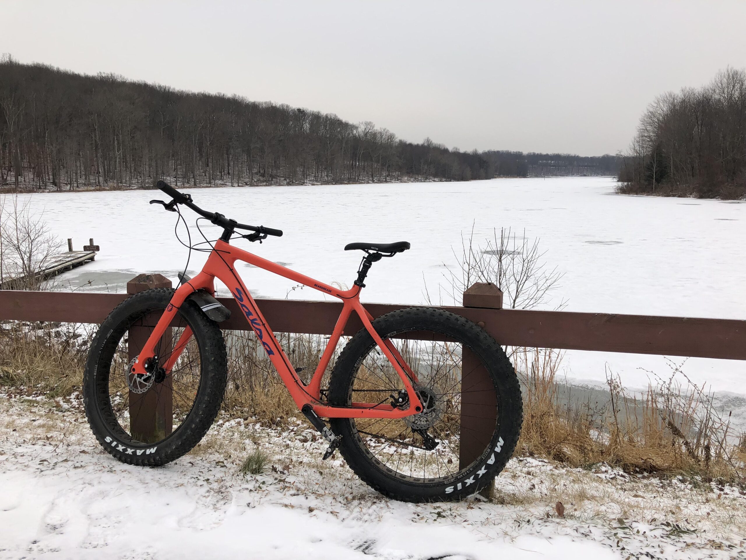 Salsa Beargrease: An orange fat bike rests against a wooden railing by a frozen lake, surrounded by snow and leafless trees. The icy surface of the water reflects a gray sky, while a small dock can be seen in the background. The scene captures a quiet, wintry landscape.