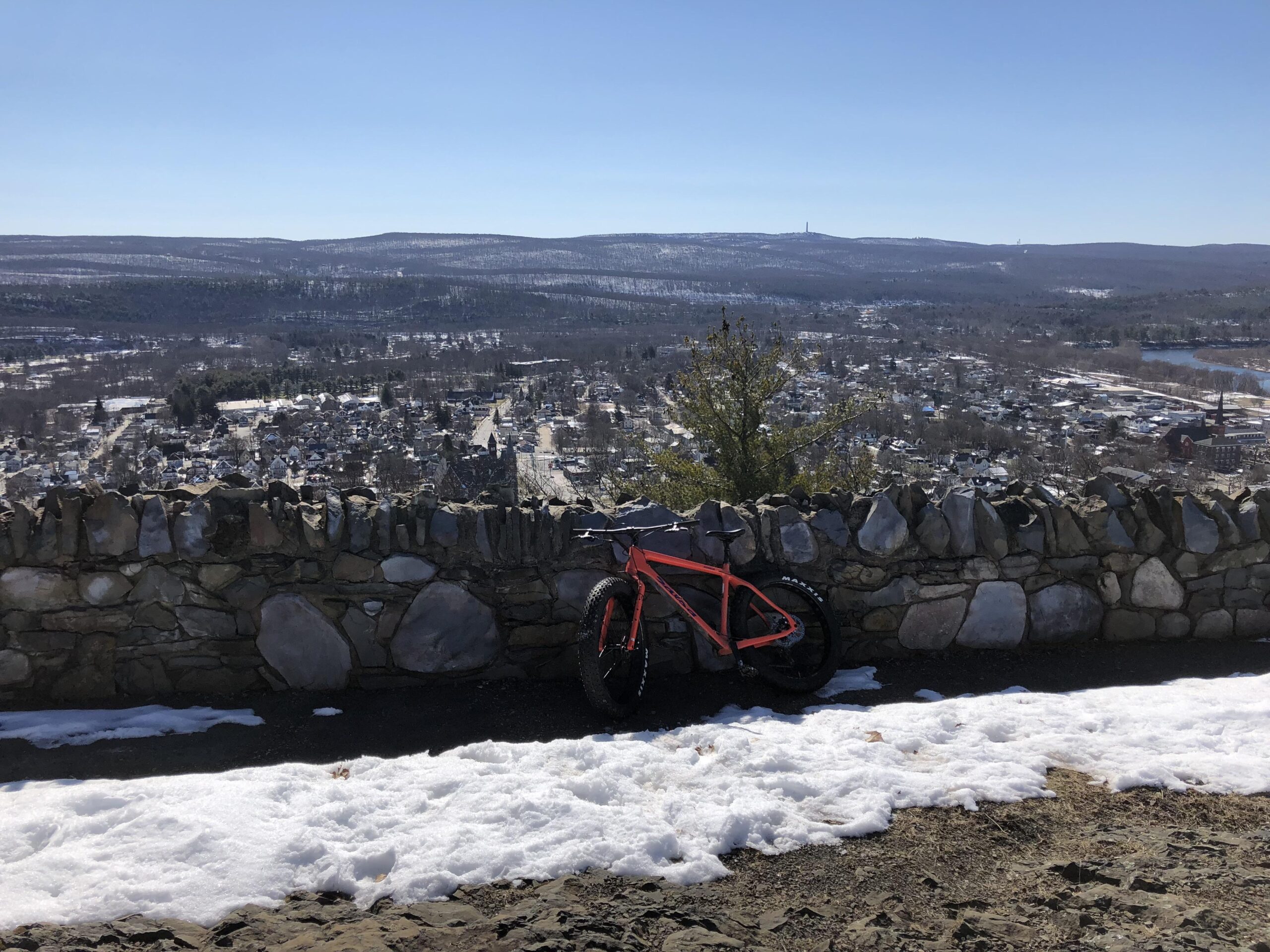 Salsa Beargrease: A vibrant orange fat bike is leaned against a stone wall overlooking a snow-covered valley and a small town below, with clear blue skies and distant mountains in the background. Snow is visible on the ground, and the landscape is a mix of white and earthy tones.
