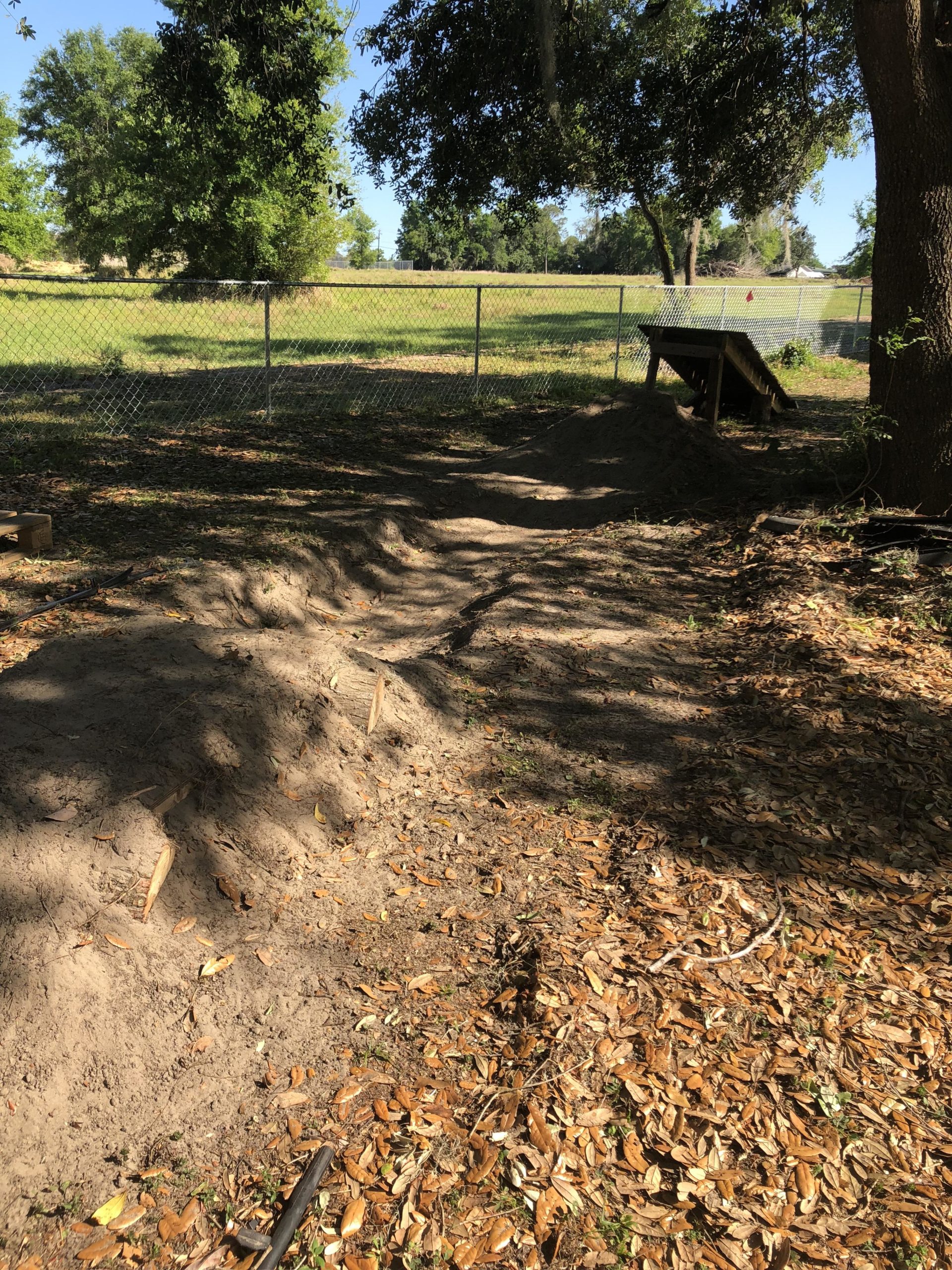 A dirt pathway with scattered leaves, leading through a fenced area with trees. In the background, a grassy field is visible beyond the chain-link fence, and a small wooden structure is partially seen on the right side of the image. Thunder Creek mountain bike trail.